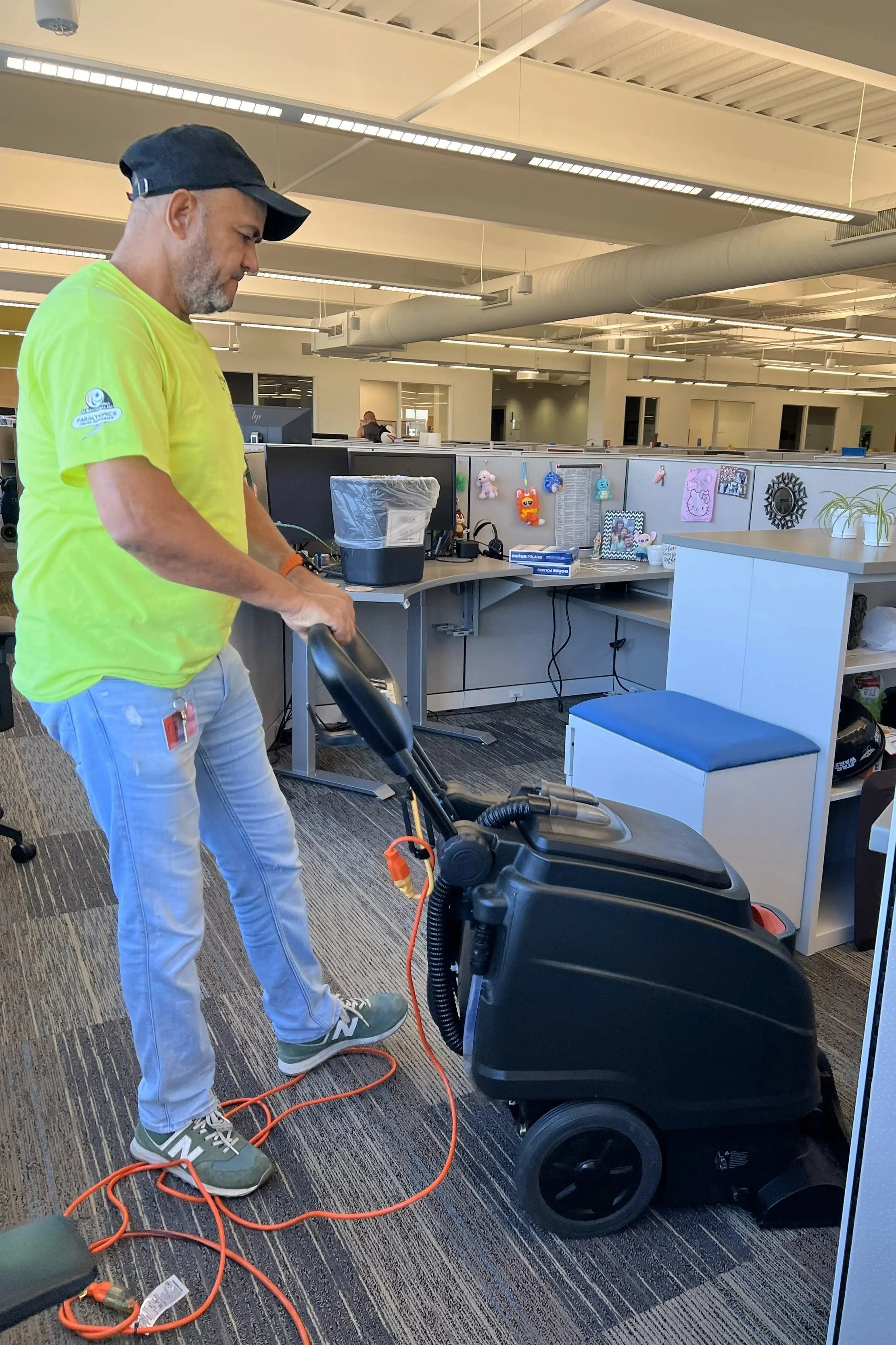 A man wearing a neon yellow shirt, gray pants, and green sneakers, is vacuuming an office carpet with a black commercial vacuum cleaner. The office has cubicles, computers, and decorations, with a ceiling featuring exposed ducts and lighting.