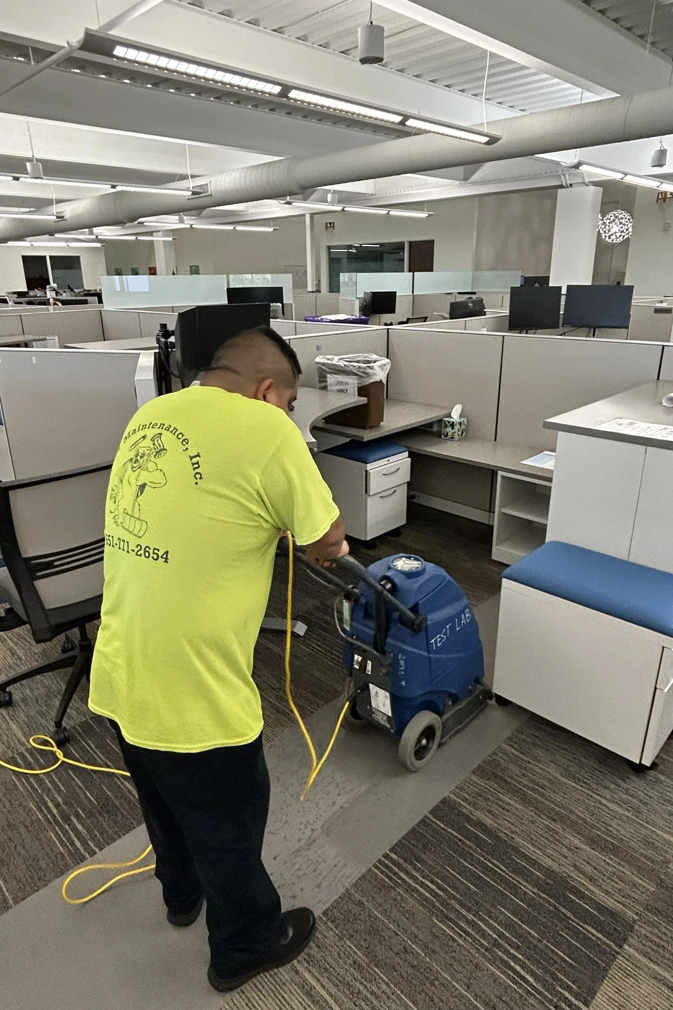 A worker in a yellow shirt cleaning the office carpet with a blue carpet cleaning machine.