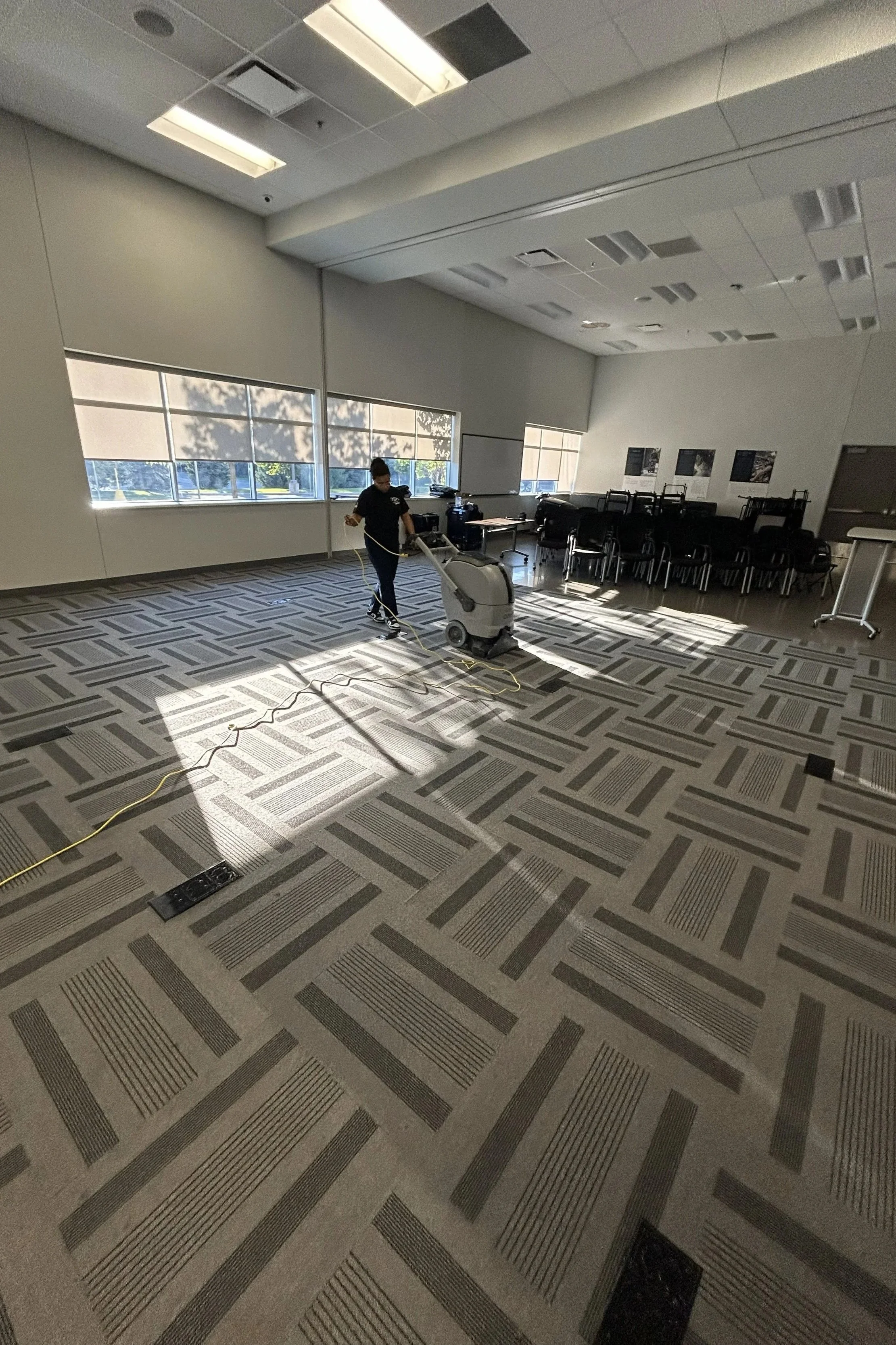 Person cleaning the empty conference or classroom with a floor cleaning machine, sunlight coming through large windows, chairs stacked at the back, and some pictures on the wall.