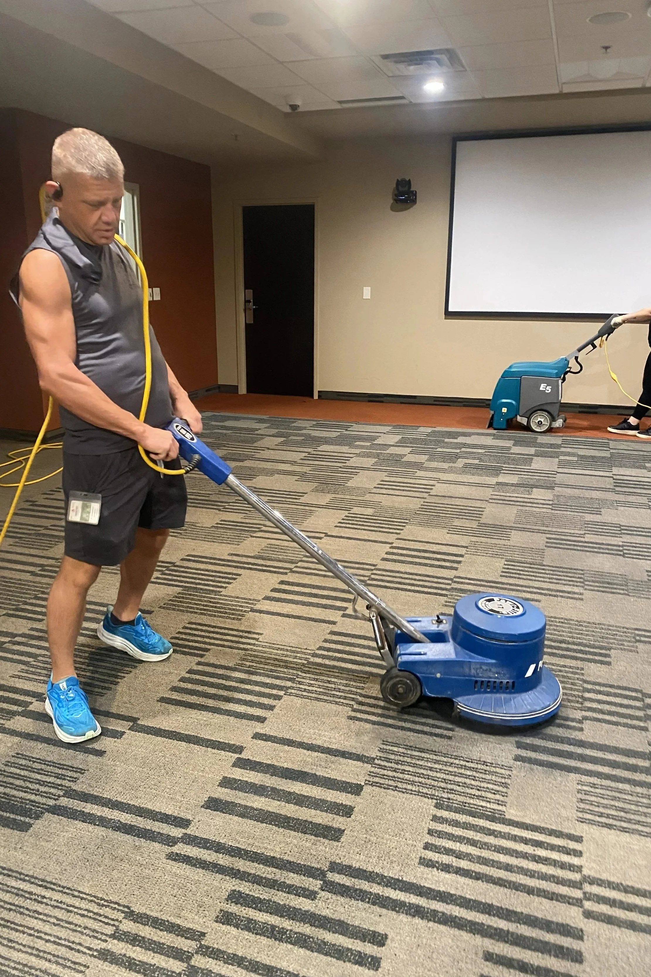 A man wearing athletic clothing and blue sneakers is operating a blue carpet cleaning machine in a conference room with patterned carpet and a large projector screen on the wall.