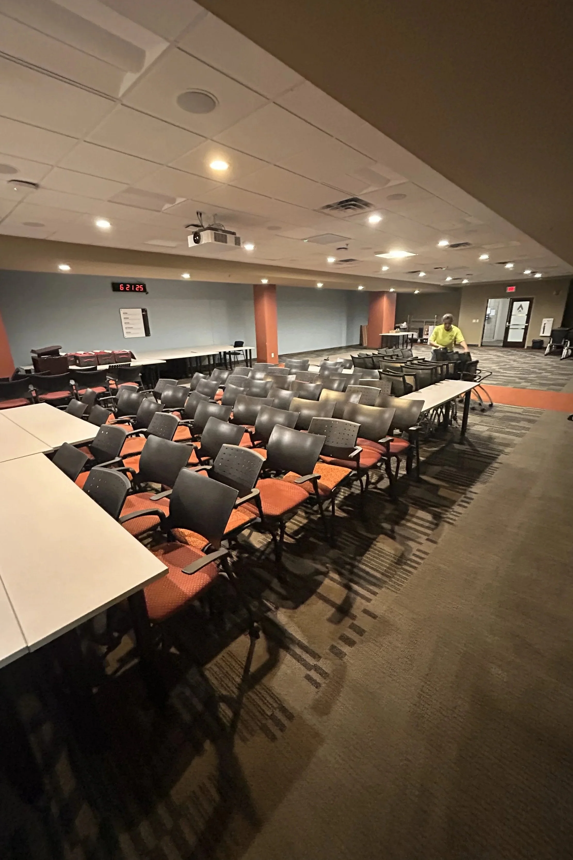 A conference room with multiple rows of black chairs with armrests and red cushions, arranged in front of long white tables. A man in a yellow shirt is standing near the tables at the back of the room. The room has a drop ceiling with recessed lighti