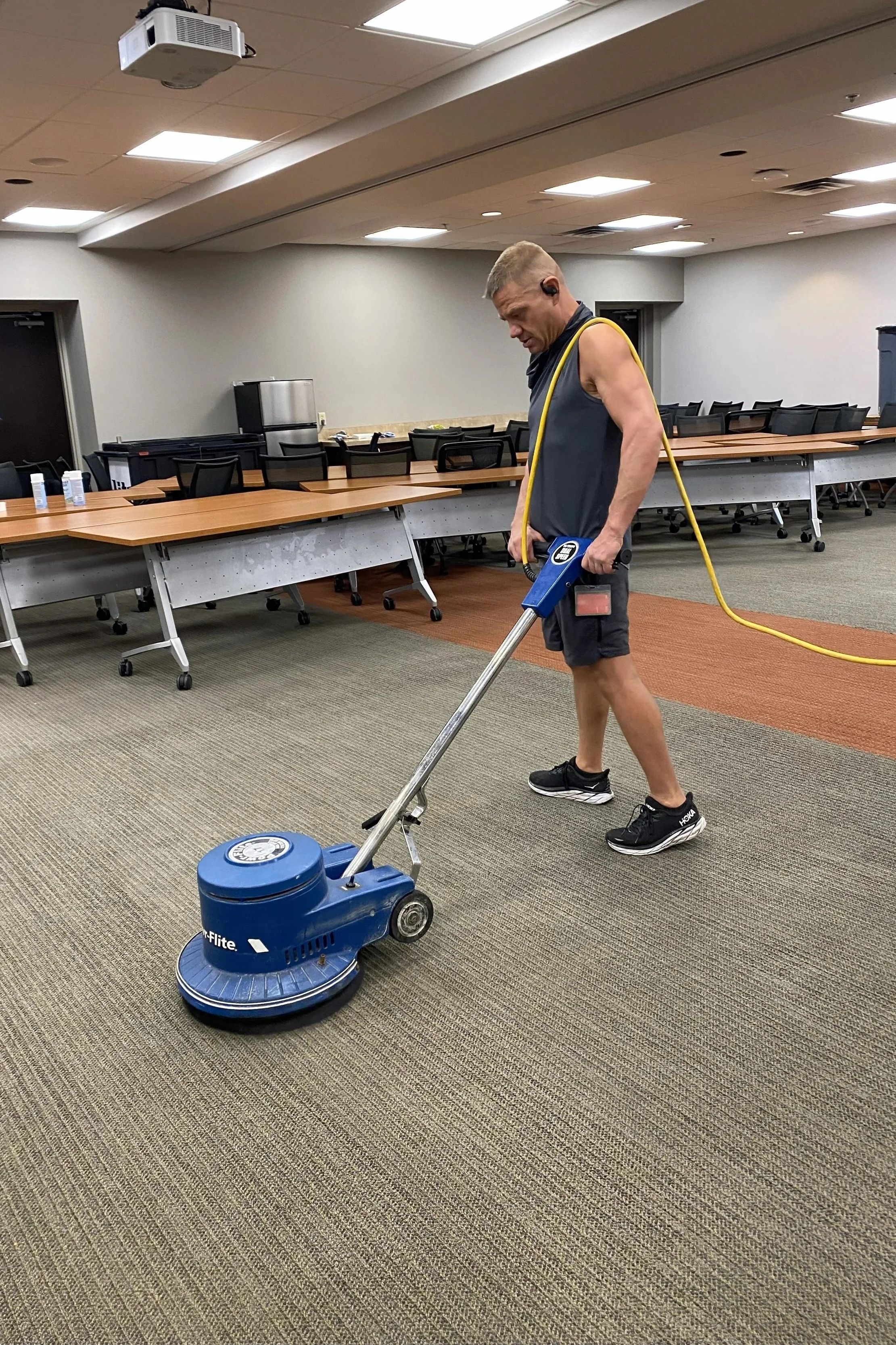 A man cleaning the carpet of a conference room with a blue industrial floor scrubber. The room has long tables and black chairs, with some bottles of sanitizer on the tables and a fridge in the corner.