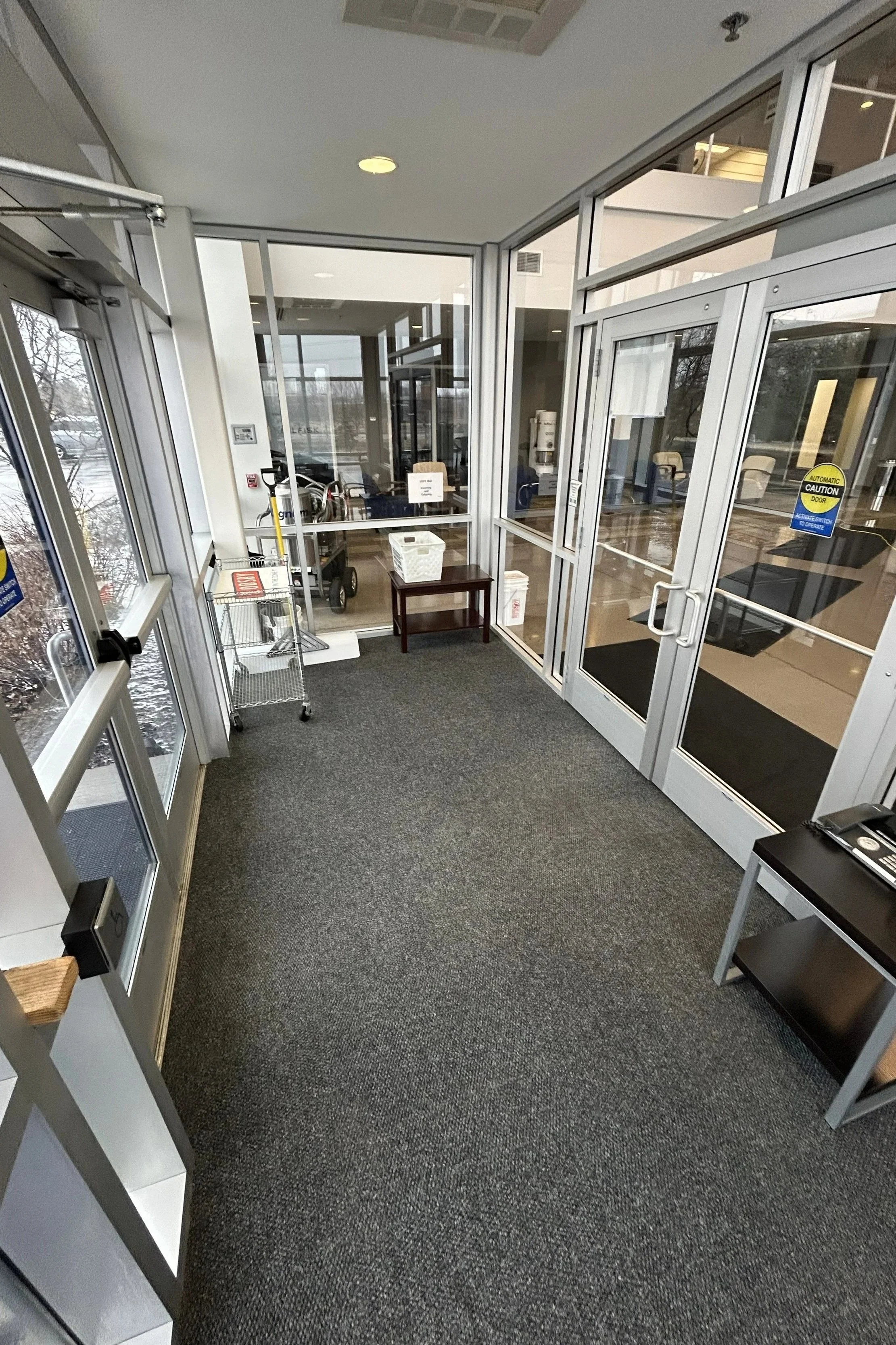 Entrance area of a building with glass doors and windows, gray carpet, a small table, and a shopping cart inside.