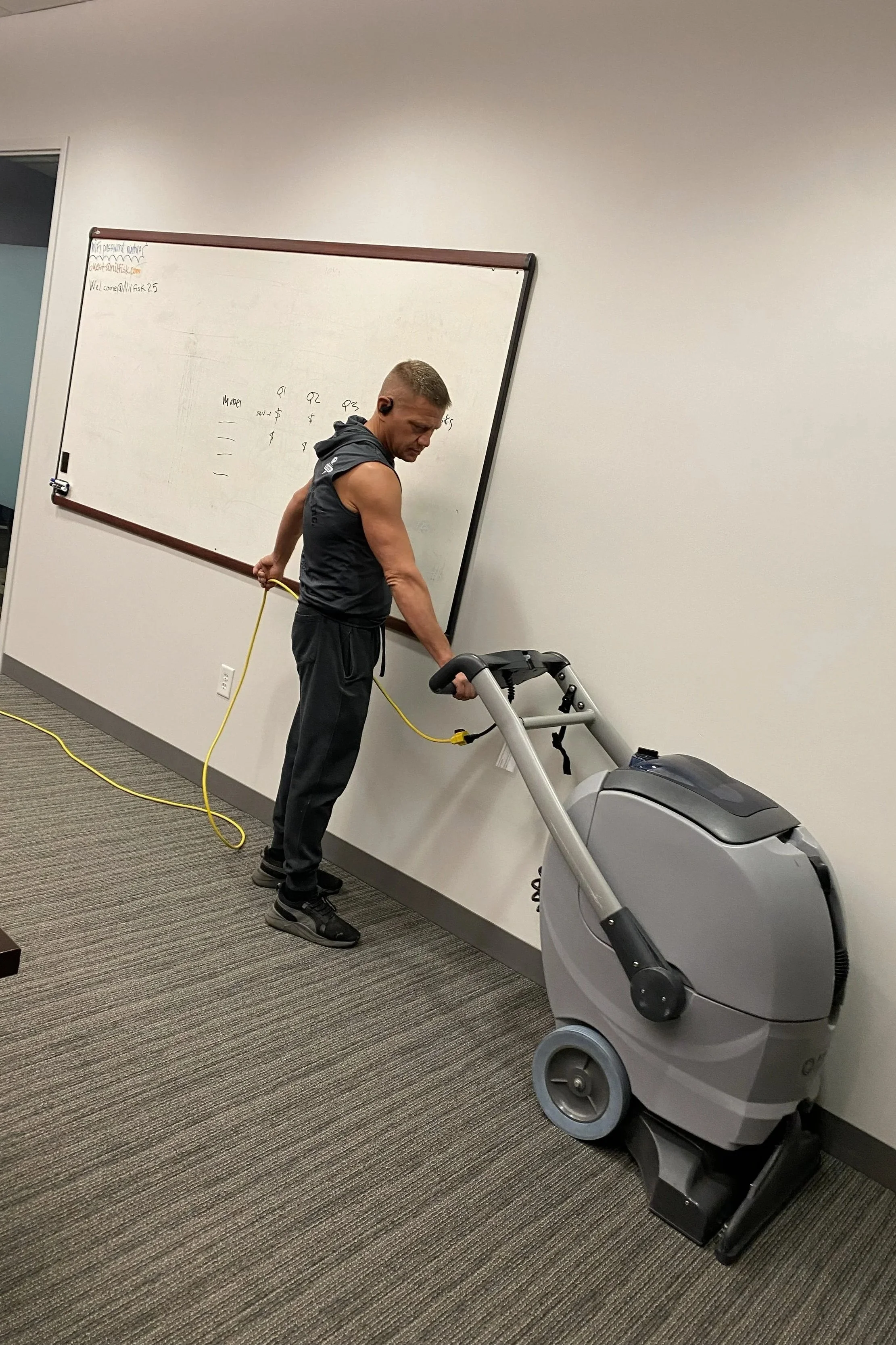 Man pushing an industrial carpet cleaning machine in an office room, with a whiteboard in the background.