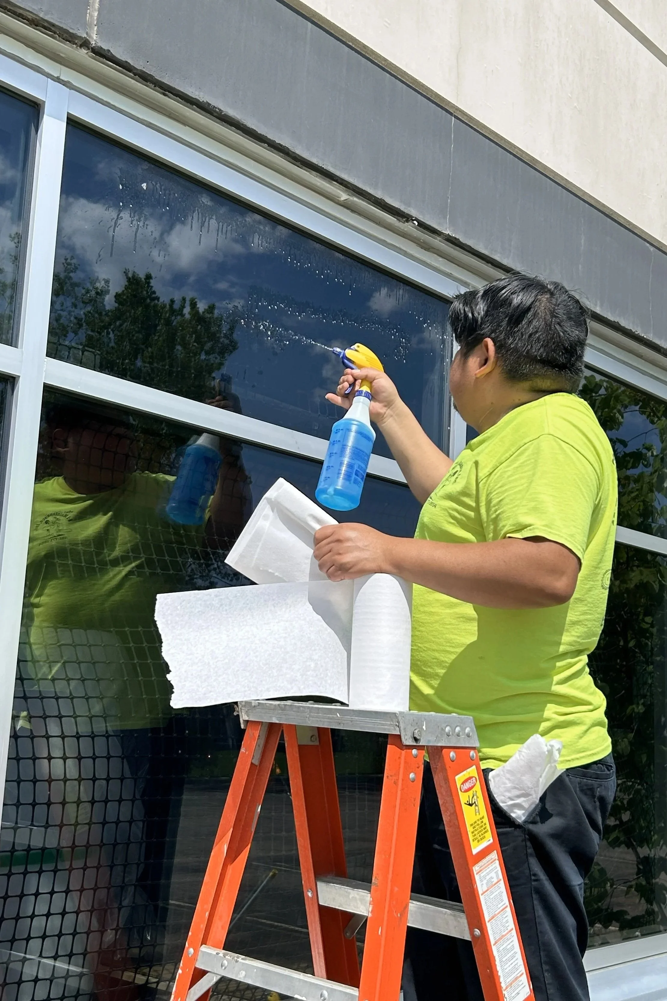 A man in a yellow shirt cleaning a large window from the outside with a spray bottle and cloth, standing on an orange ladder.