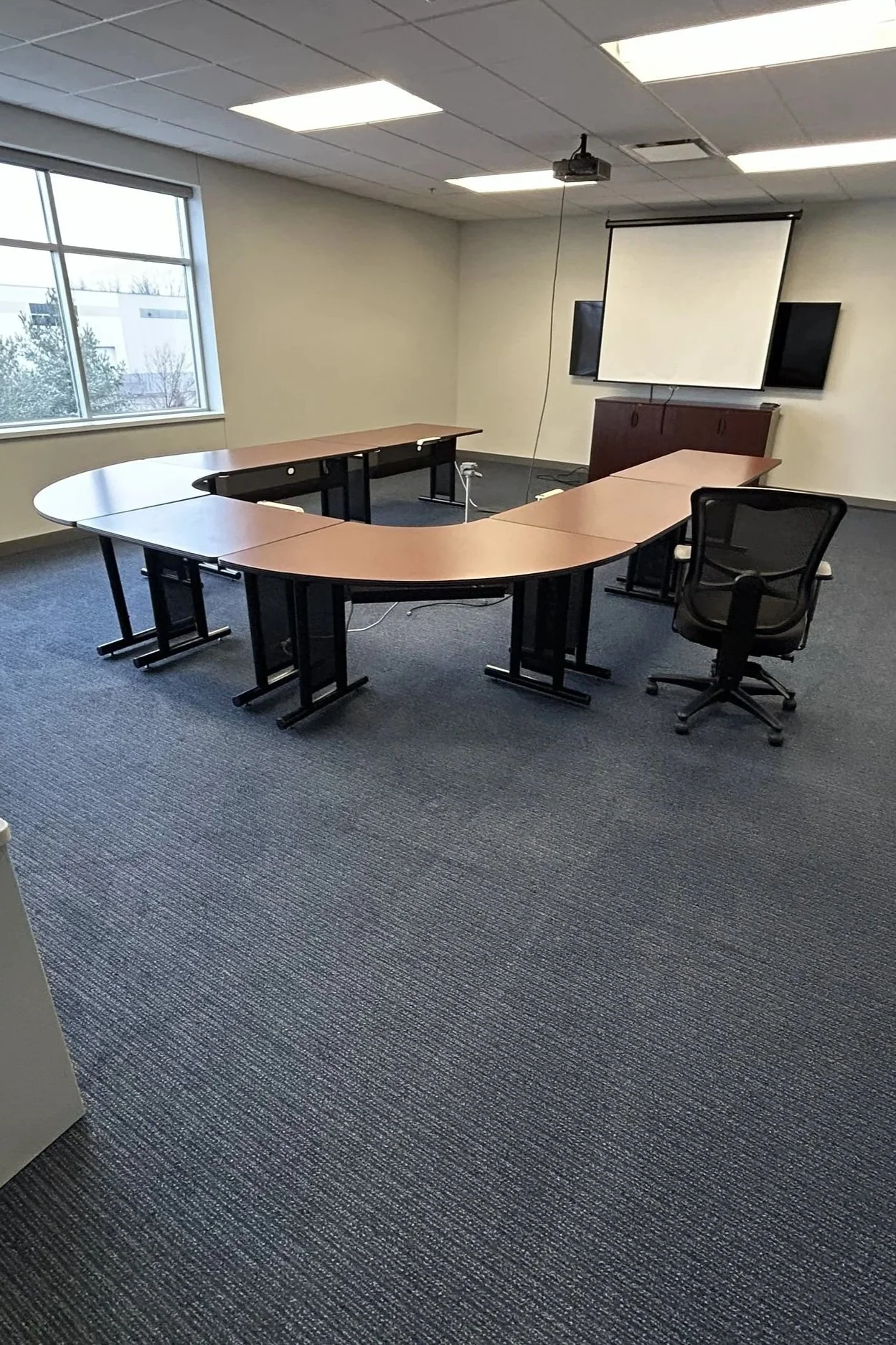 Empty conference room with U-shaped table, black office chair, large window on the left, a ceiling-mounted projector, and a pull-down screen, with minimal decorations.