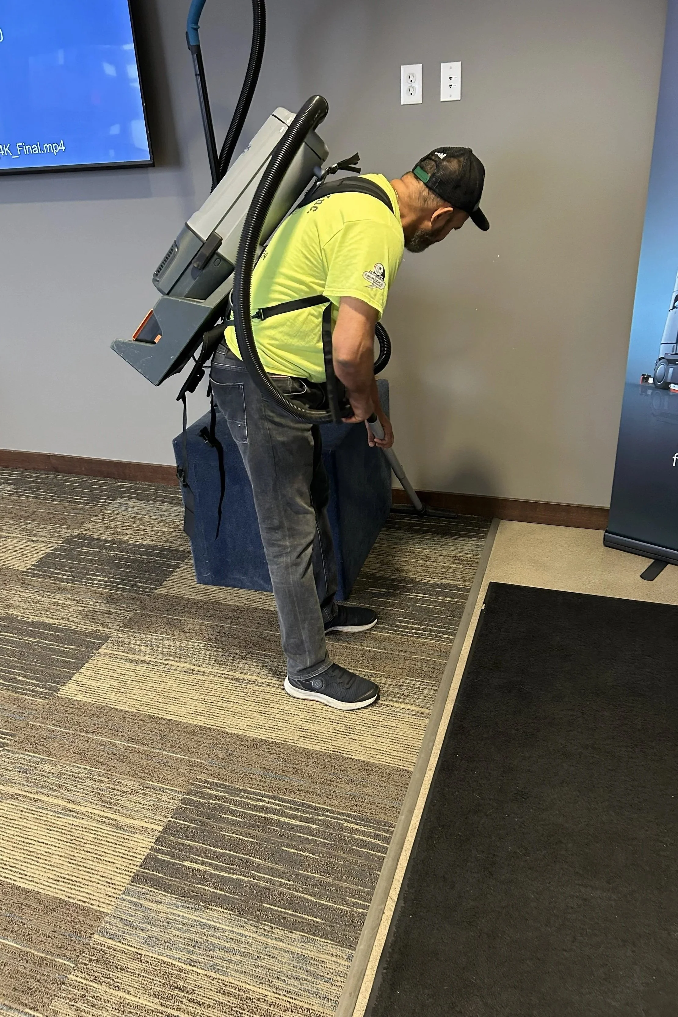 A man wearing a black cap, yellow shirt, and dark pants is using a carpet extractor machine to clean the carpet near a wall with electrical outlets. The machine is worn on his back, and he is bent over as he works.