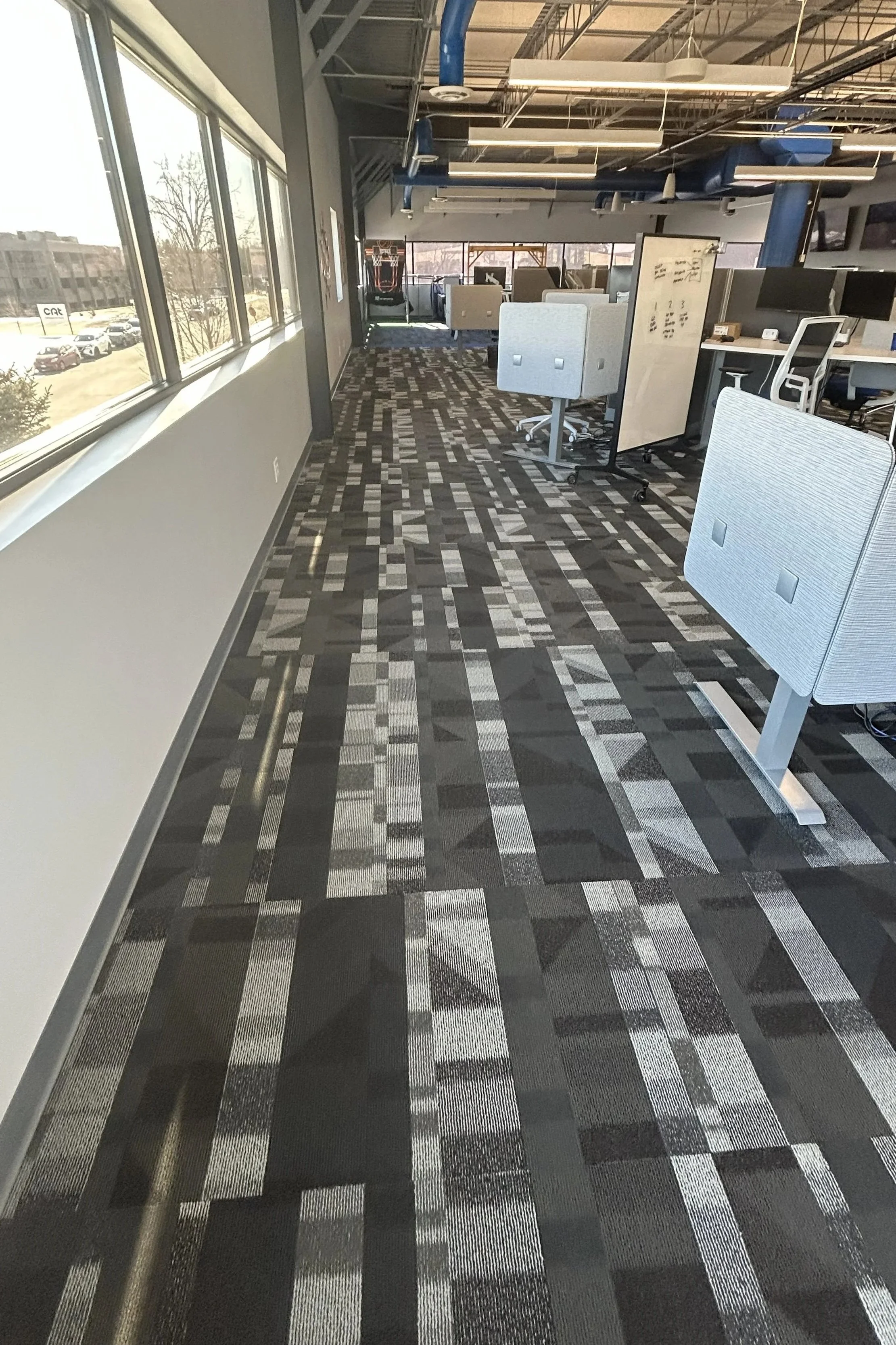 An office with large windows on the left, patterned carpeting, white rolling chairs, a whiteboard, and work desks with office chairs under a ceiling with exposed ducts and lights.