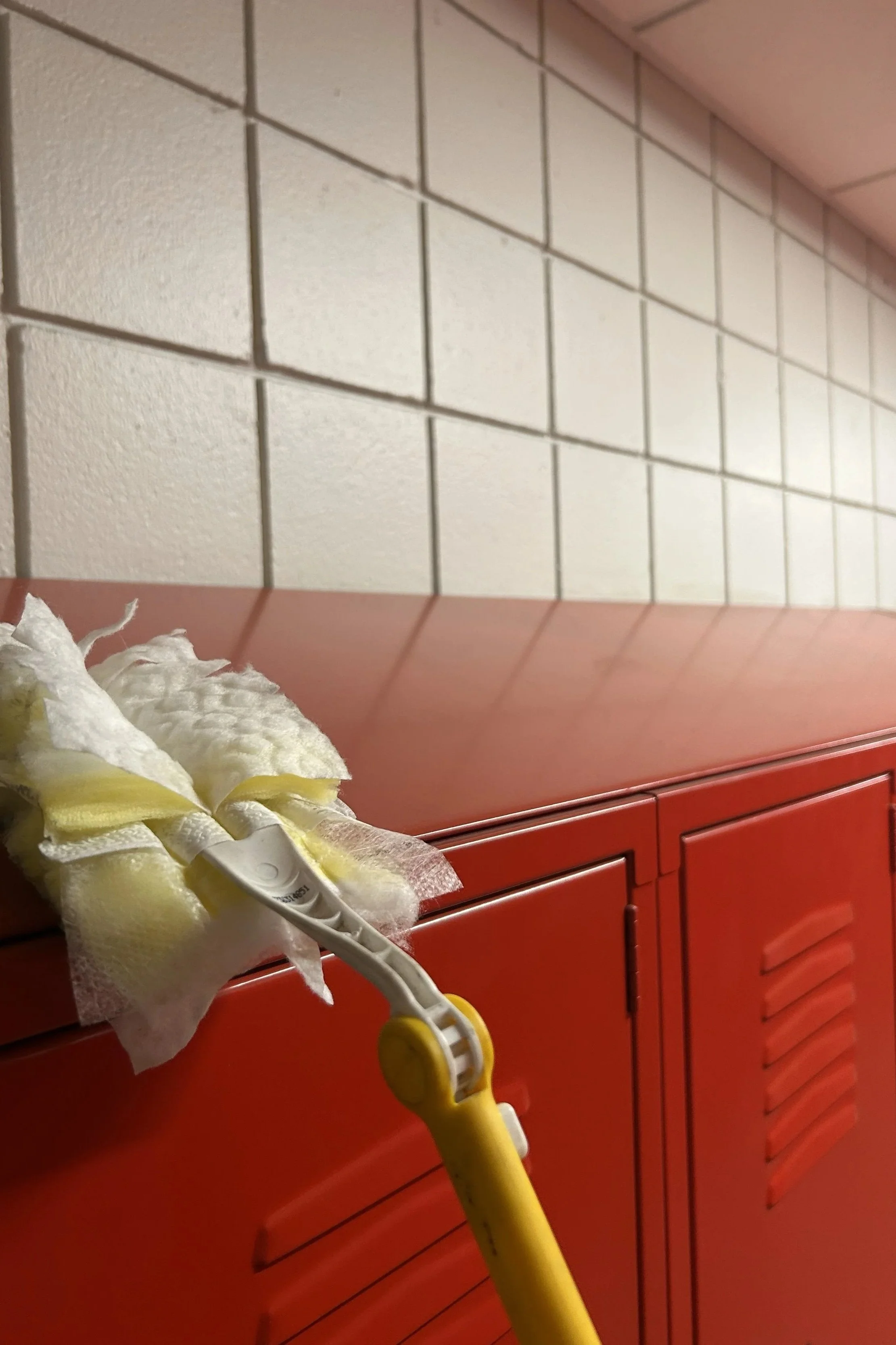 A red school locker with a mop leaning against it, in a hallway with white tiled walls.