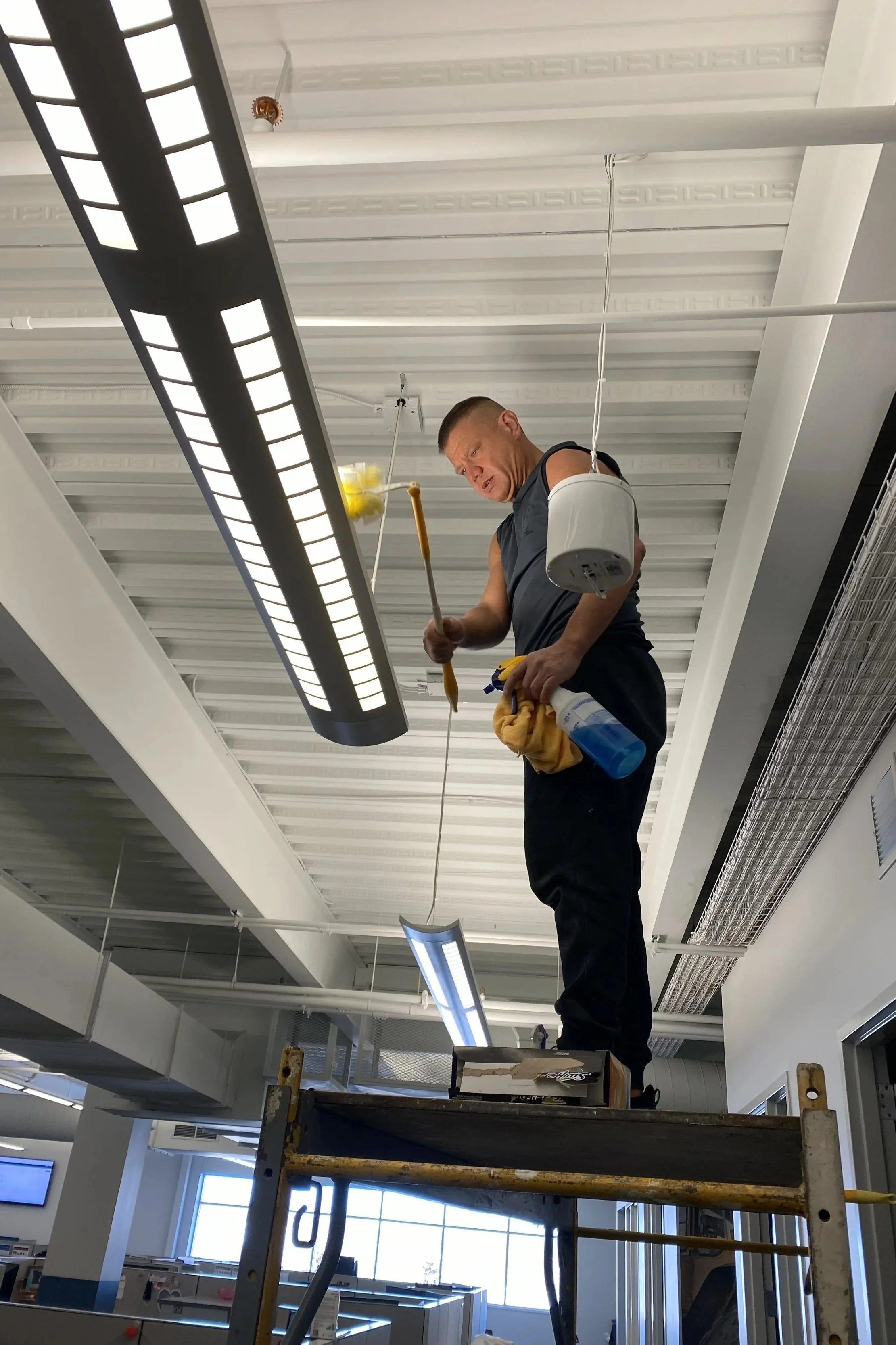 A man standing on scaffolding cleaning ceiling lights in an office with a yellow cloth and spray bottle.