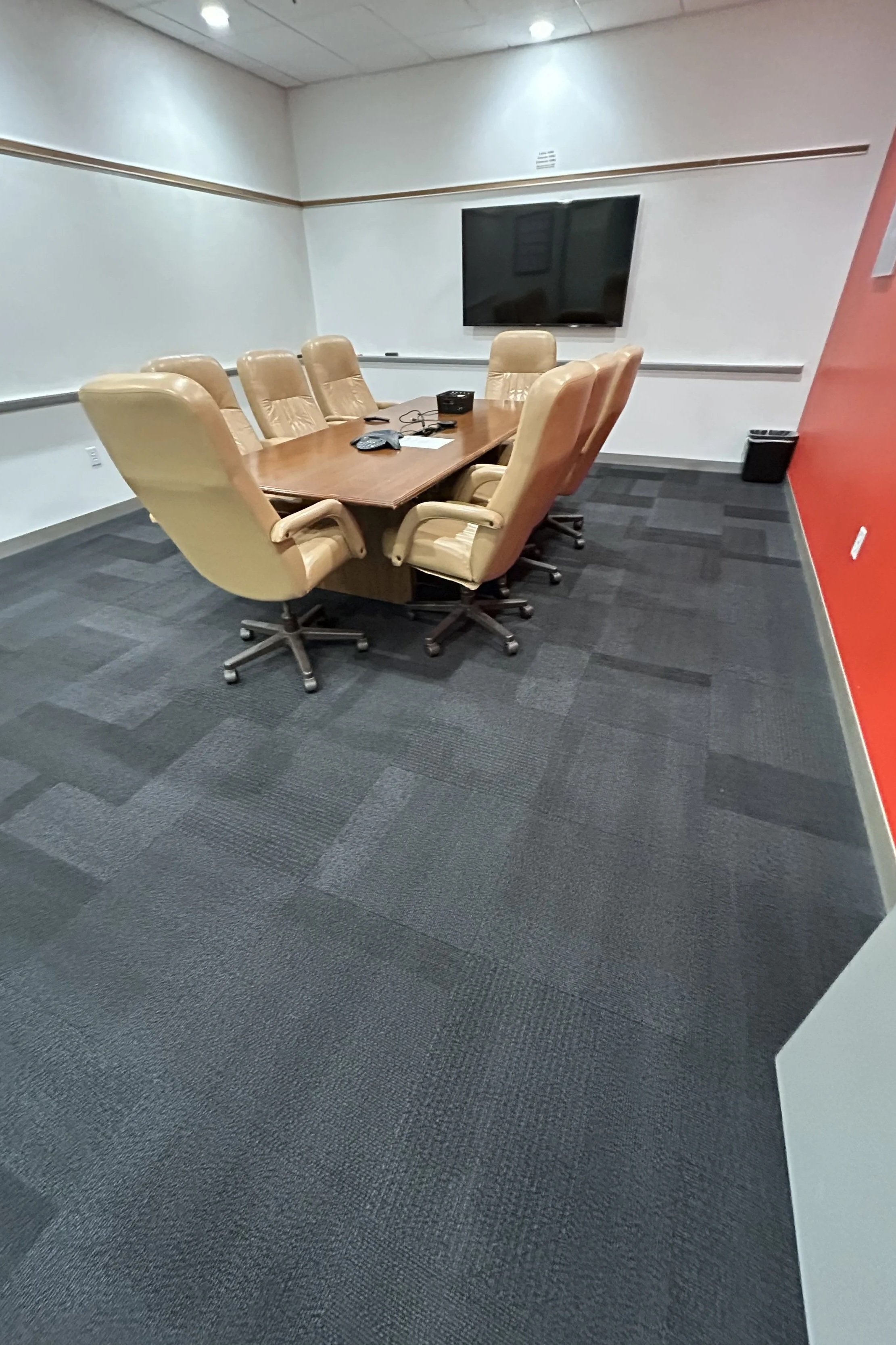Empty conference room with a wooden table surrounded by nine beige office chairs, a flat-screen TV on the back wall, and a black trash bin in the corner.