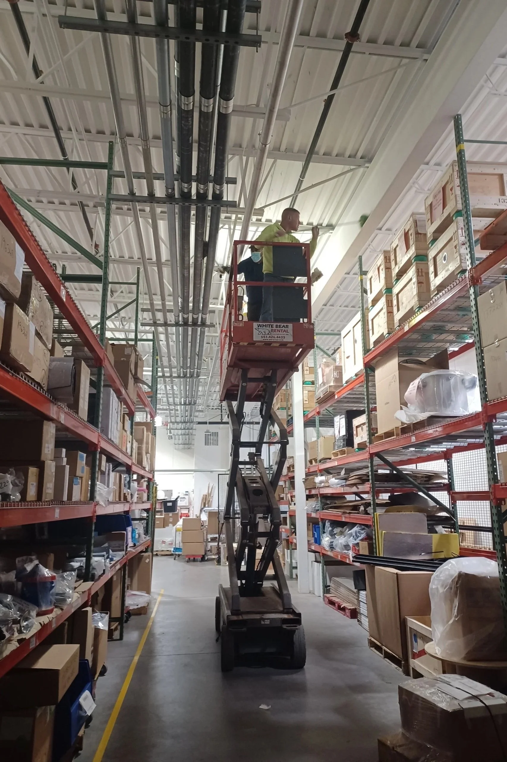 Two workers on a scissor lift working on the ceiling of a storage aisle filled with boxes and supplies.