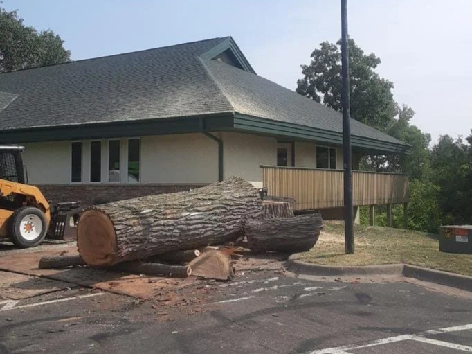 Large fallen tree trunk in front of a house with a sloped roof, part of a construction or cleanup scene with equipment nearby.