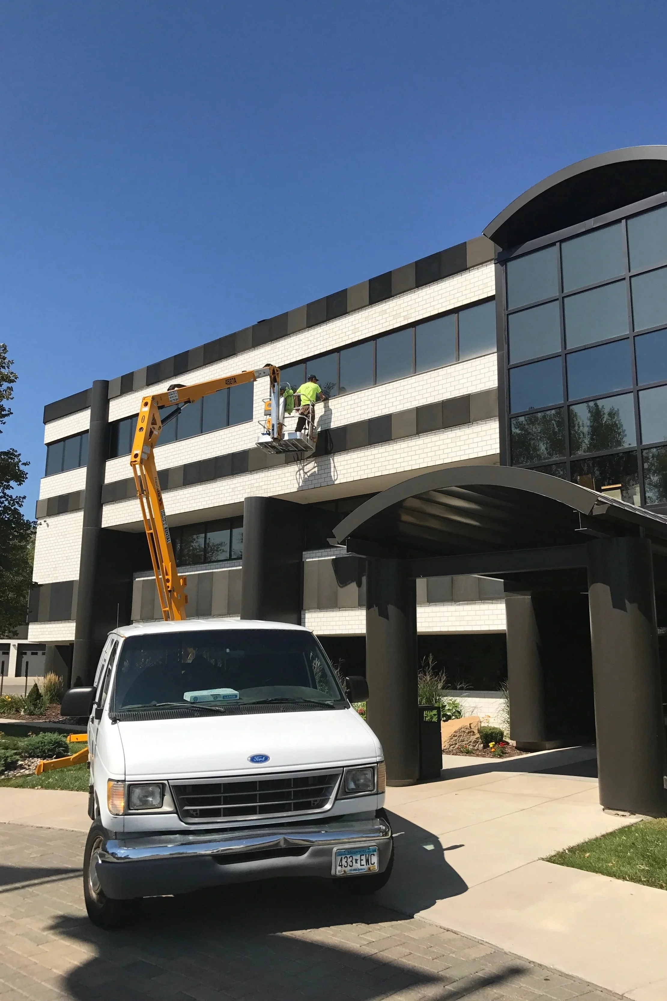 Two workers in neon safety shirts are on a lift working on the exterior of a modern office building with black and white tile and large glass windows.