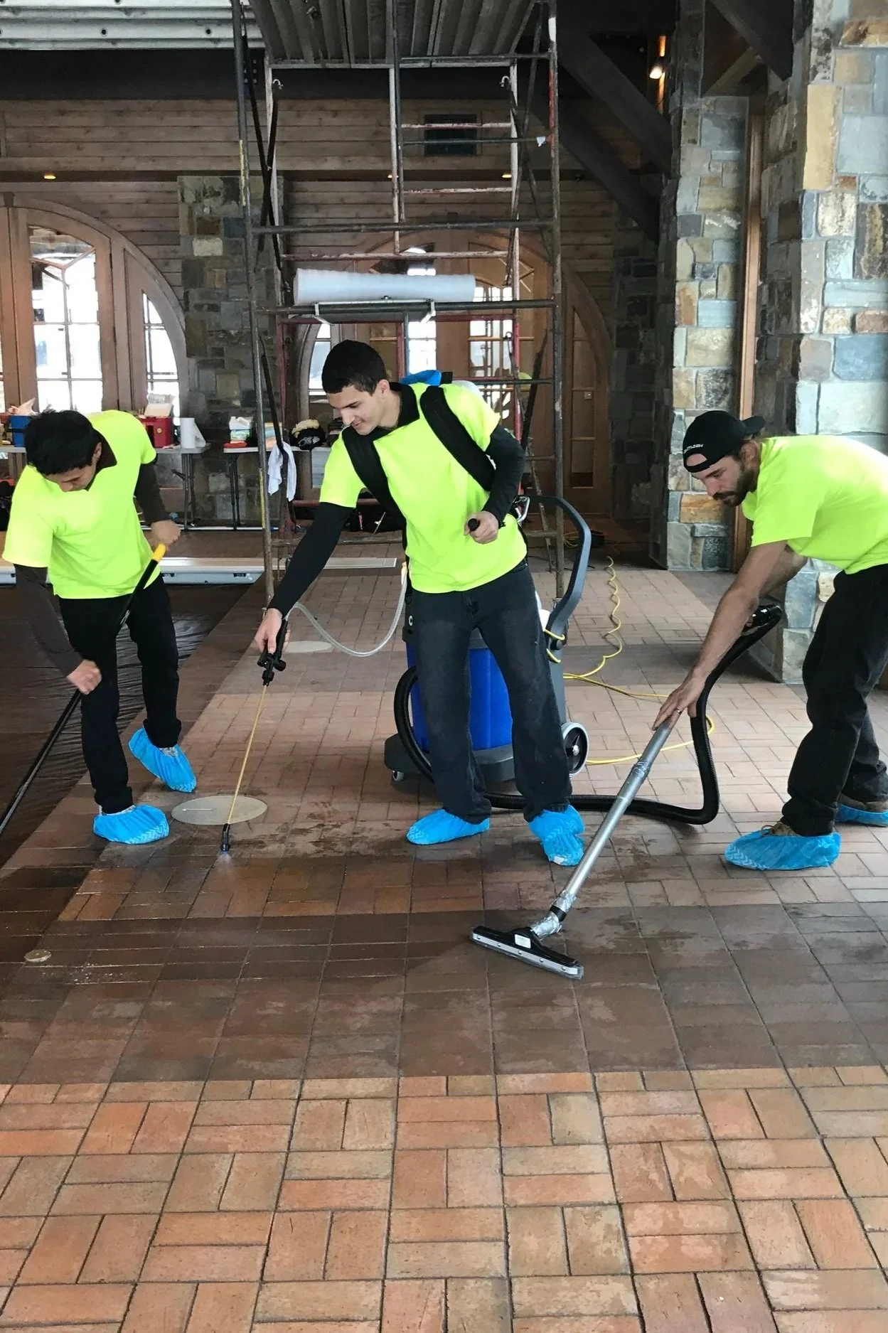 Three workers wearing safety shoe covers and fluorescent shirts are cleaning and scrubbing a brick and tile floor inside a building with stone walls and wooden ceiling, using various cleaning tools and equipment.