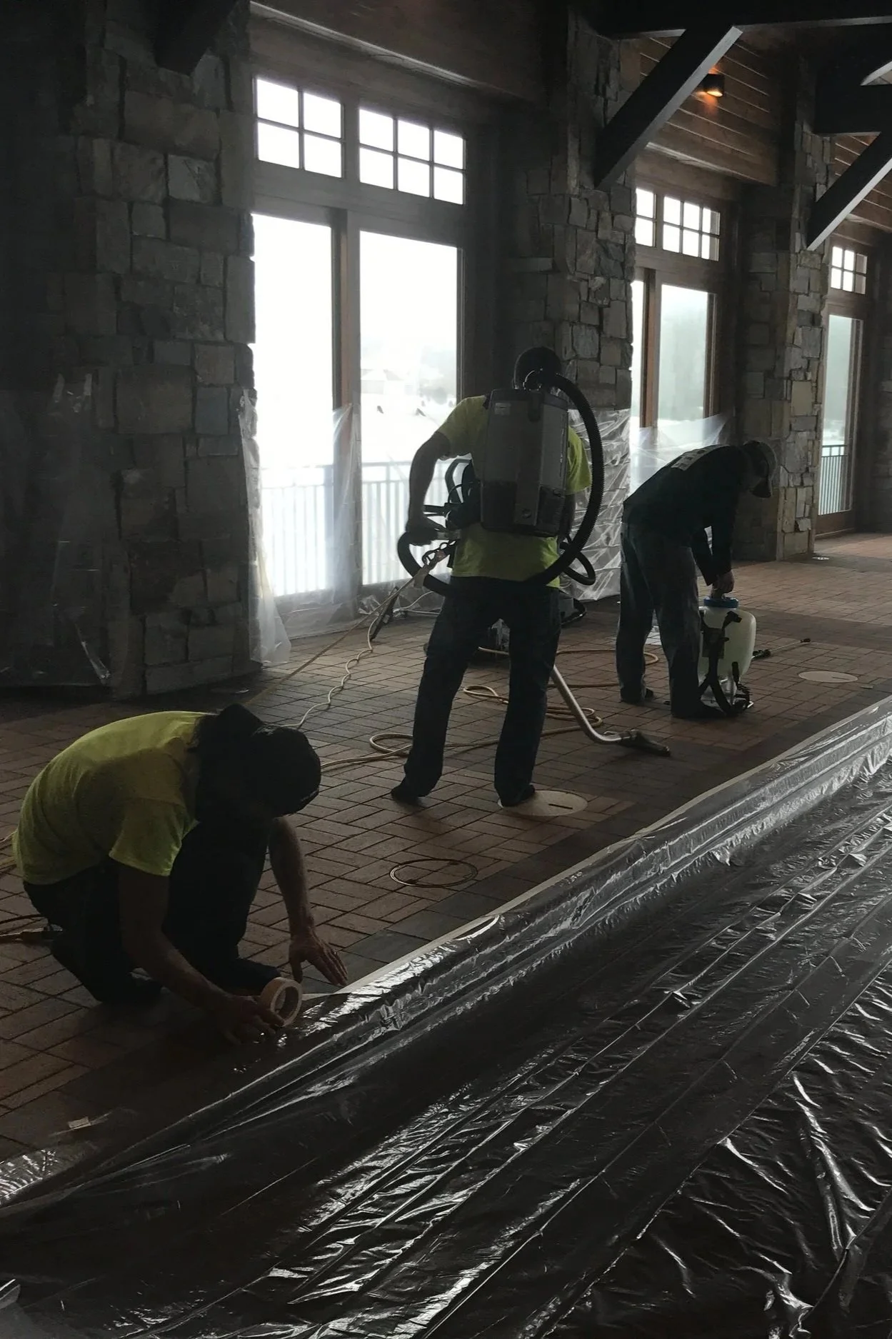 Workers installing or repairing a plastic sheeting or vapor barrier on a brick floor inside a building with large windows and stone walls.