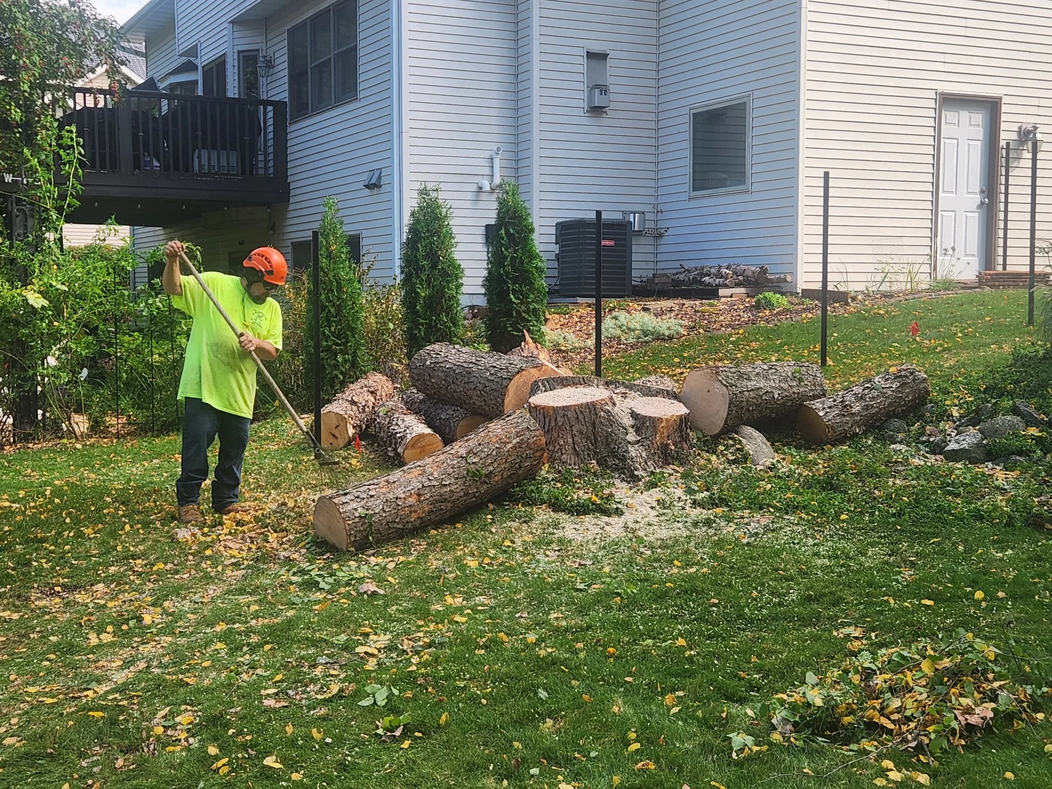 A person wearing a neon green shirt, jeans, boots, and an orange helmet is chopping wood in a backyard with a log and several cut logs on the ground. The yard has green grass with fallen leaves. A house with white siding, a deck, and an air condition