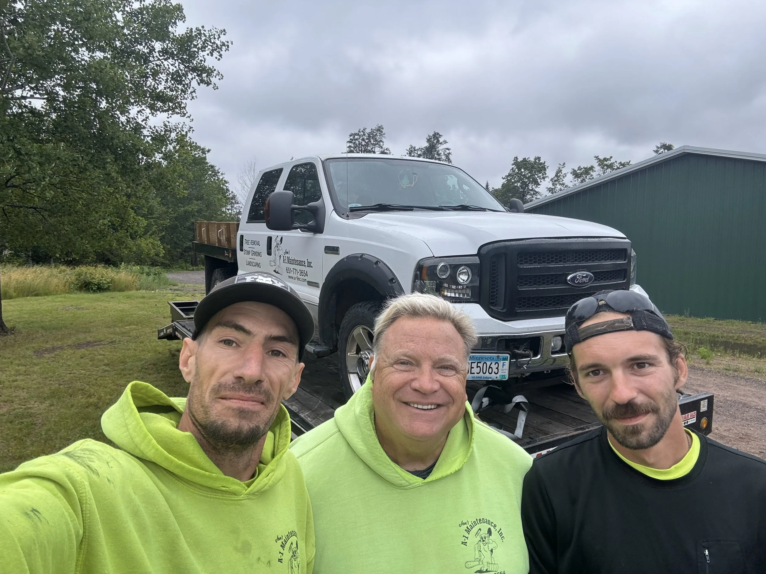 Three men taking a selfie in front of a flatbed truck with a white Ford vehicle on it, outdoors on a cloudy day surrounded by trees and a green building.