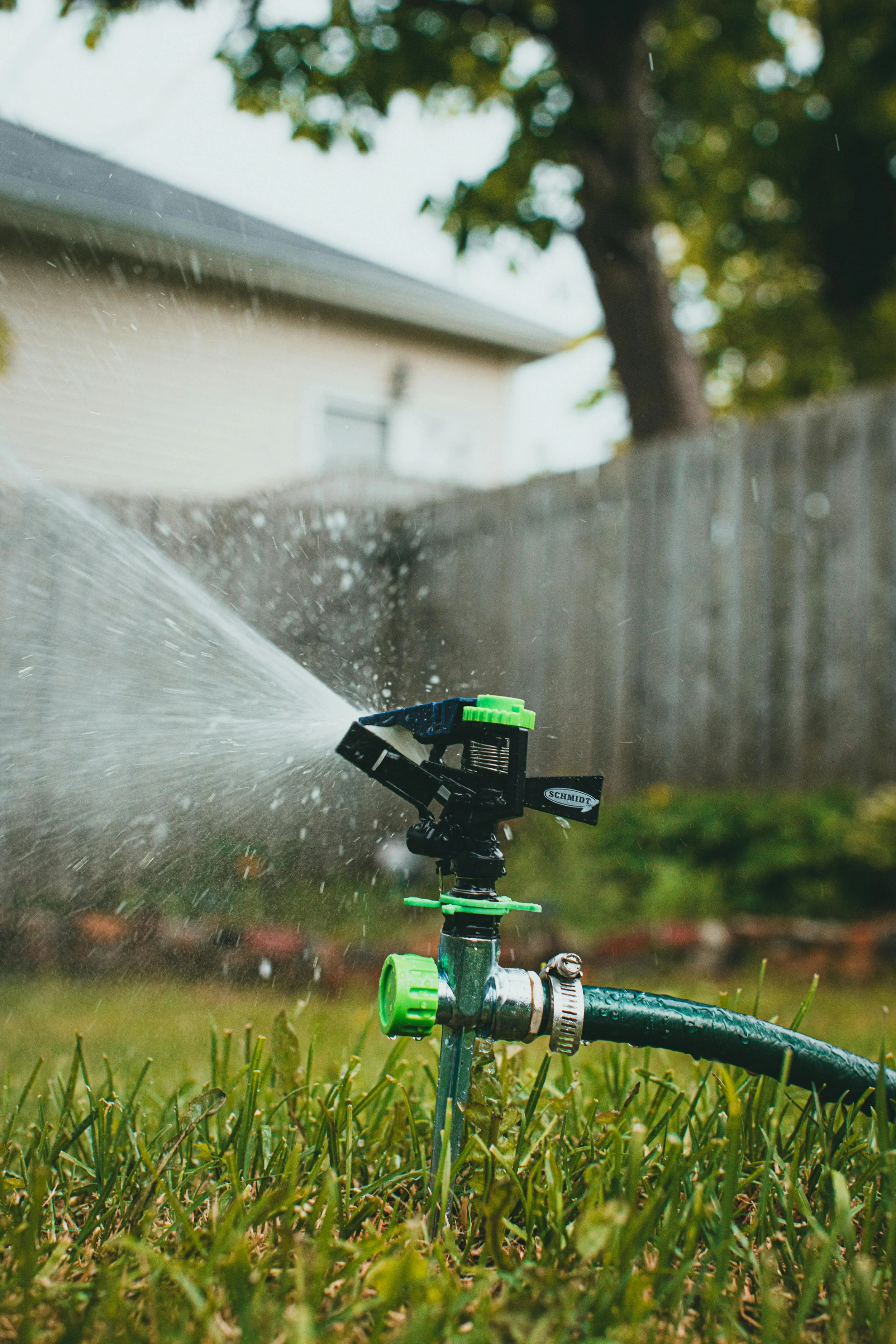 A close-up of a black and green garden sprinkler spraying water across a lawn with a house, fence, and trees in the background.