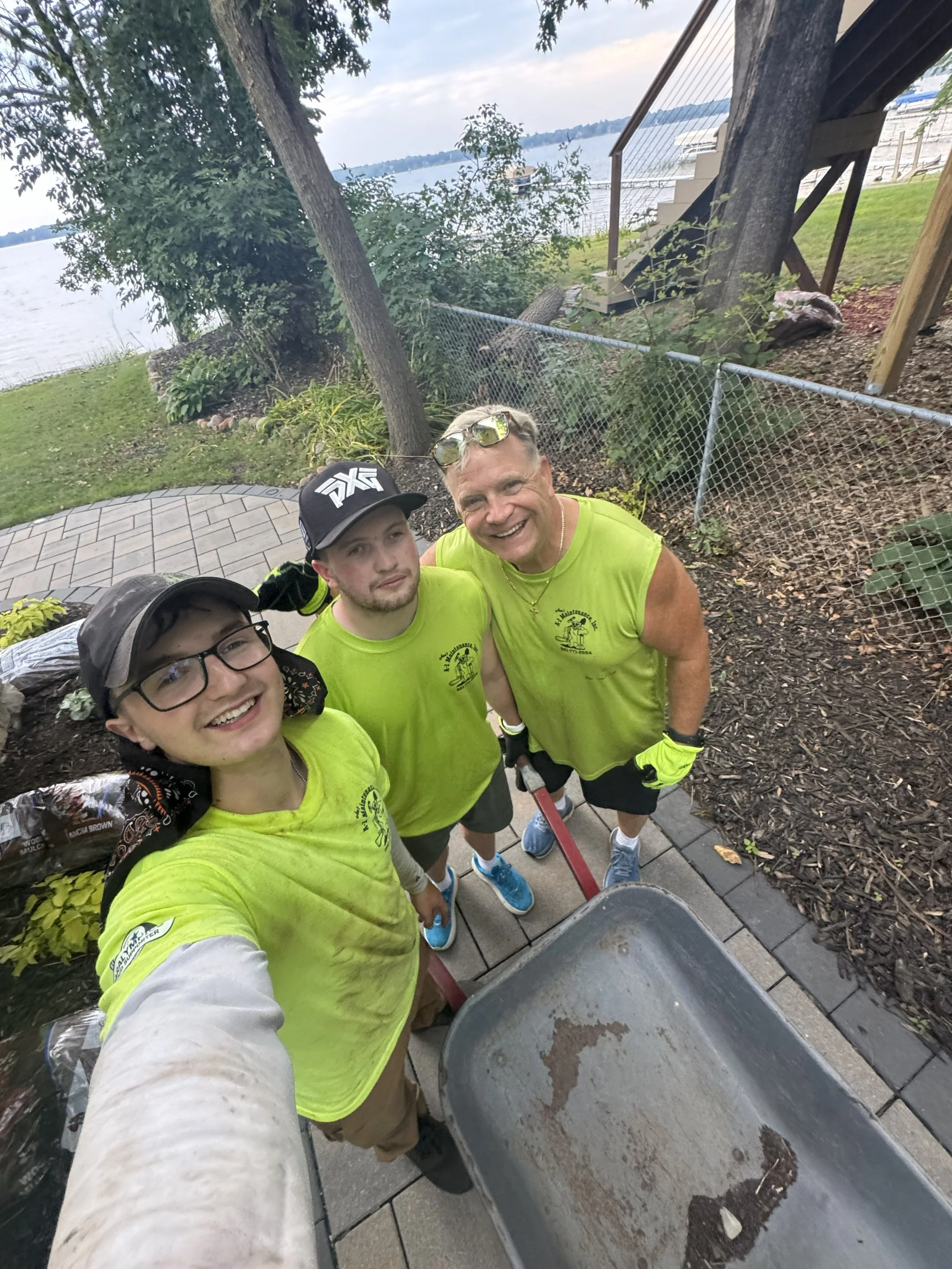 Three men taking a selfie outdoors near a waterfront, with trees, a fence, and a deck in the background. They are wearing matching neon yellow shirts and work gloves, and one is holding a wheelbarrow.