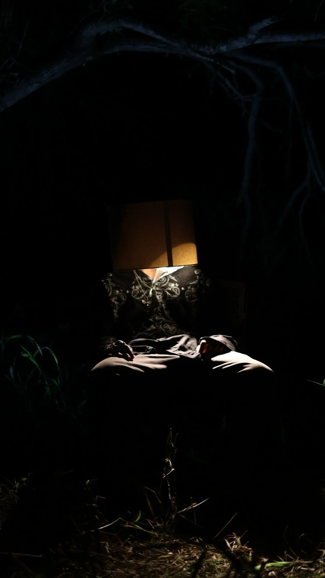 A person sitting outdoors at night, with their head replaced by a lampshade. The person is wearing dark clothing and is surrounded by greenery.