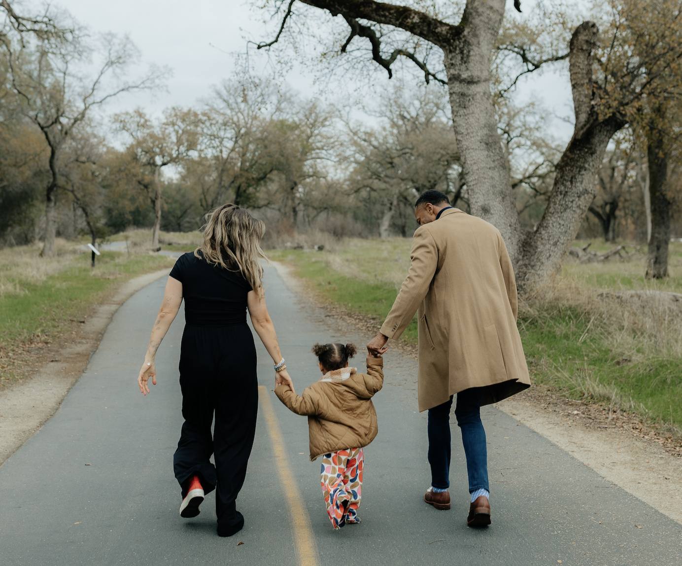 A family of three walking hand in hand on a tree-lined road during fall.