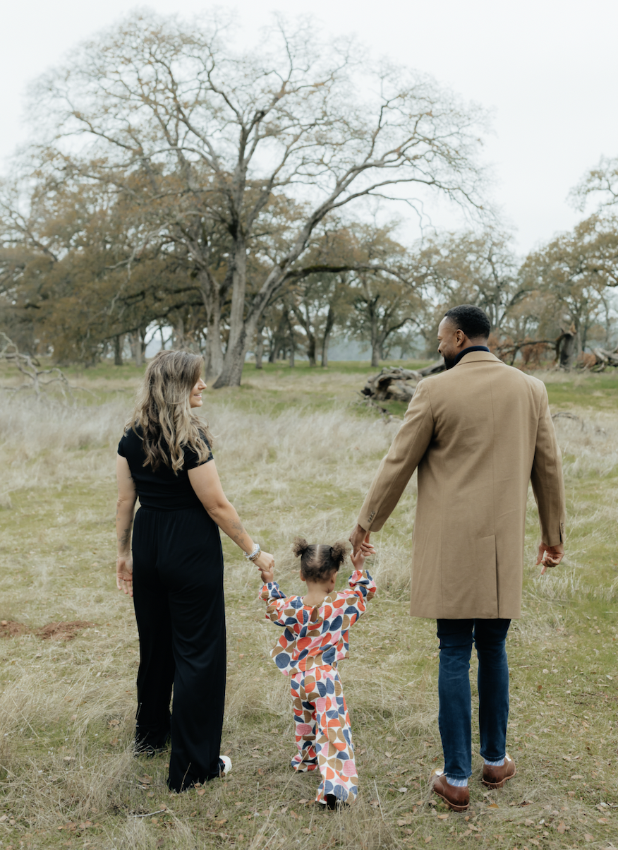 A family of three holding hands and walking in a field during autumn, with leafless trees in the background.