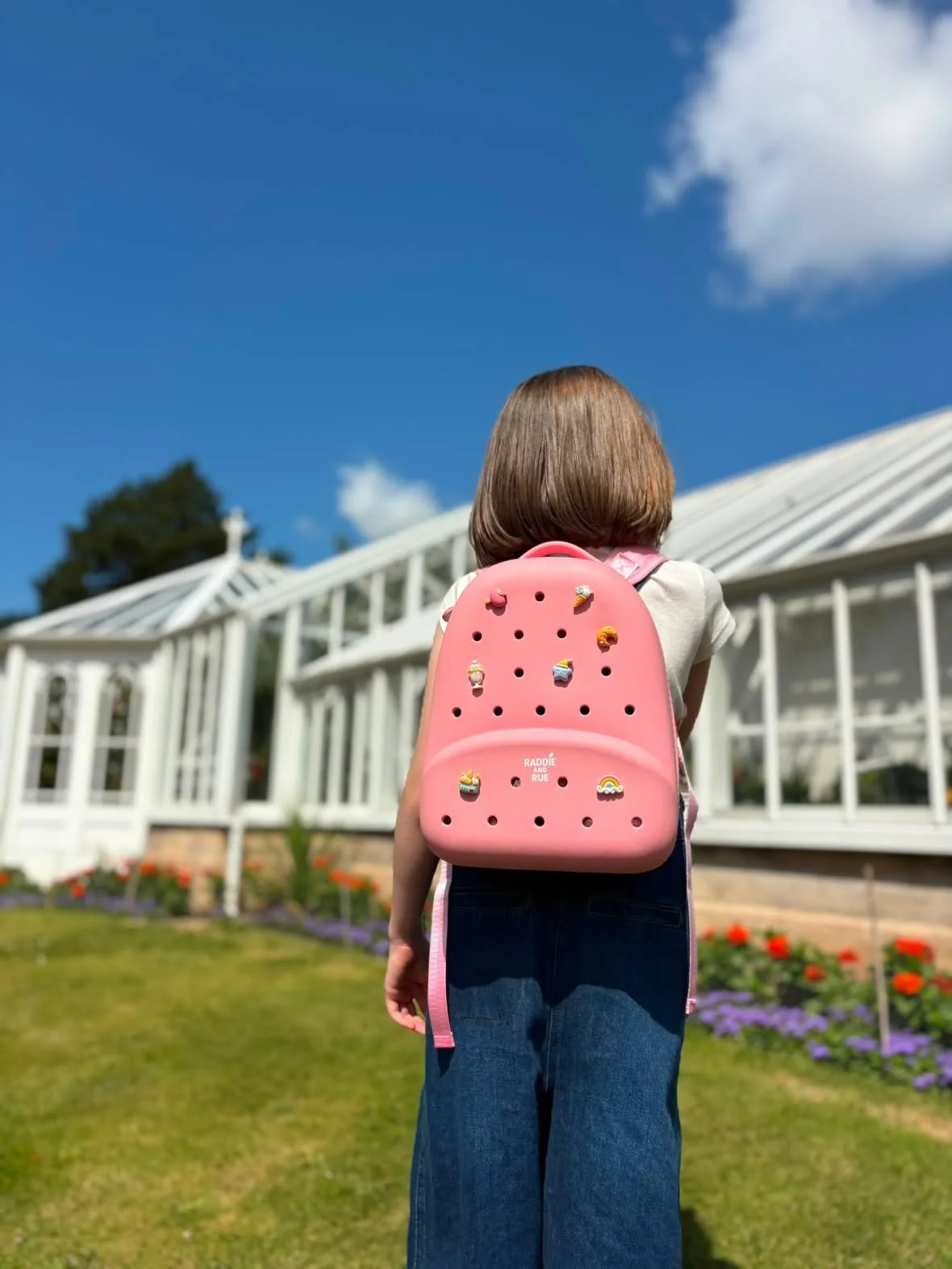 A young girl with shoulder-length hair wearing a pink backpack decorated with colorful pins, standing in front of a white greenhouse with a blue sky and clouds overhead.