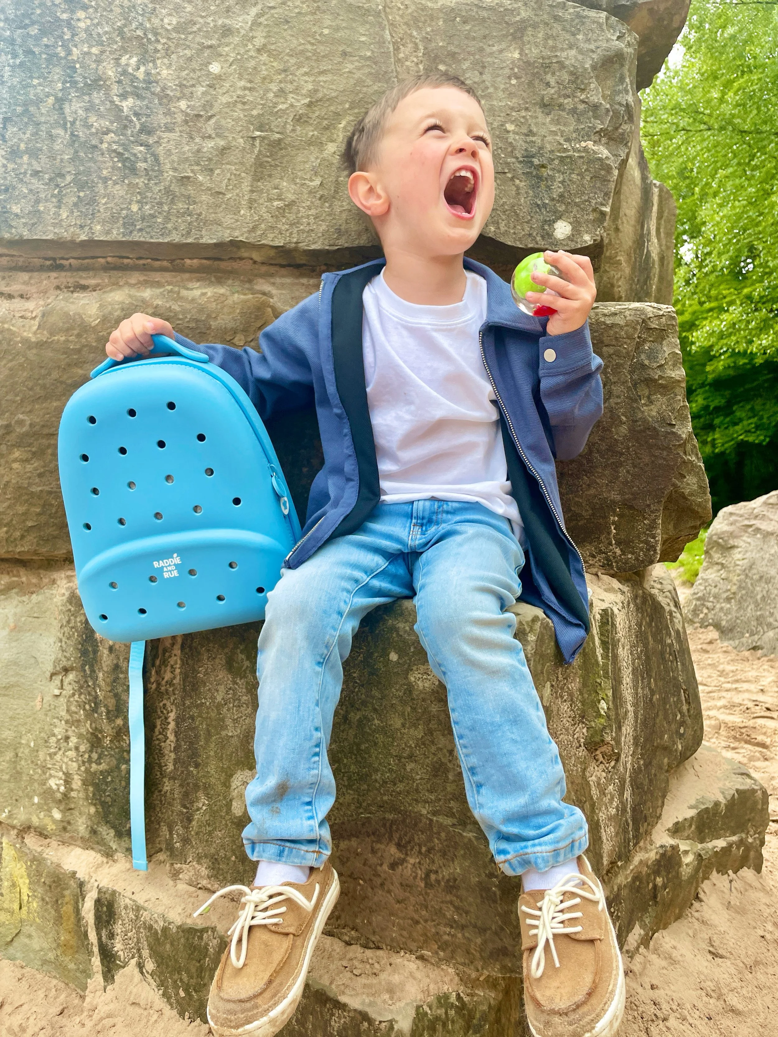 A young boy sitting on a large rock outdoors, shouting or laughing with his mouth wide open, holding a green ball in his right hand, carrying a blue backpack, wearing casual clothing including a white t-shirt, blue jeans, and tan sneakers. Behind him, there are green trees and a natural setting.