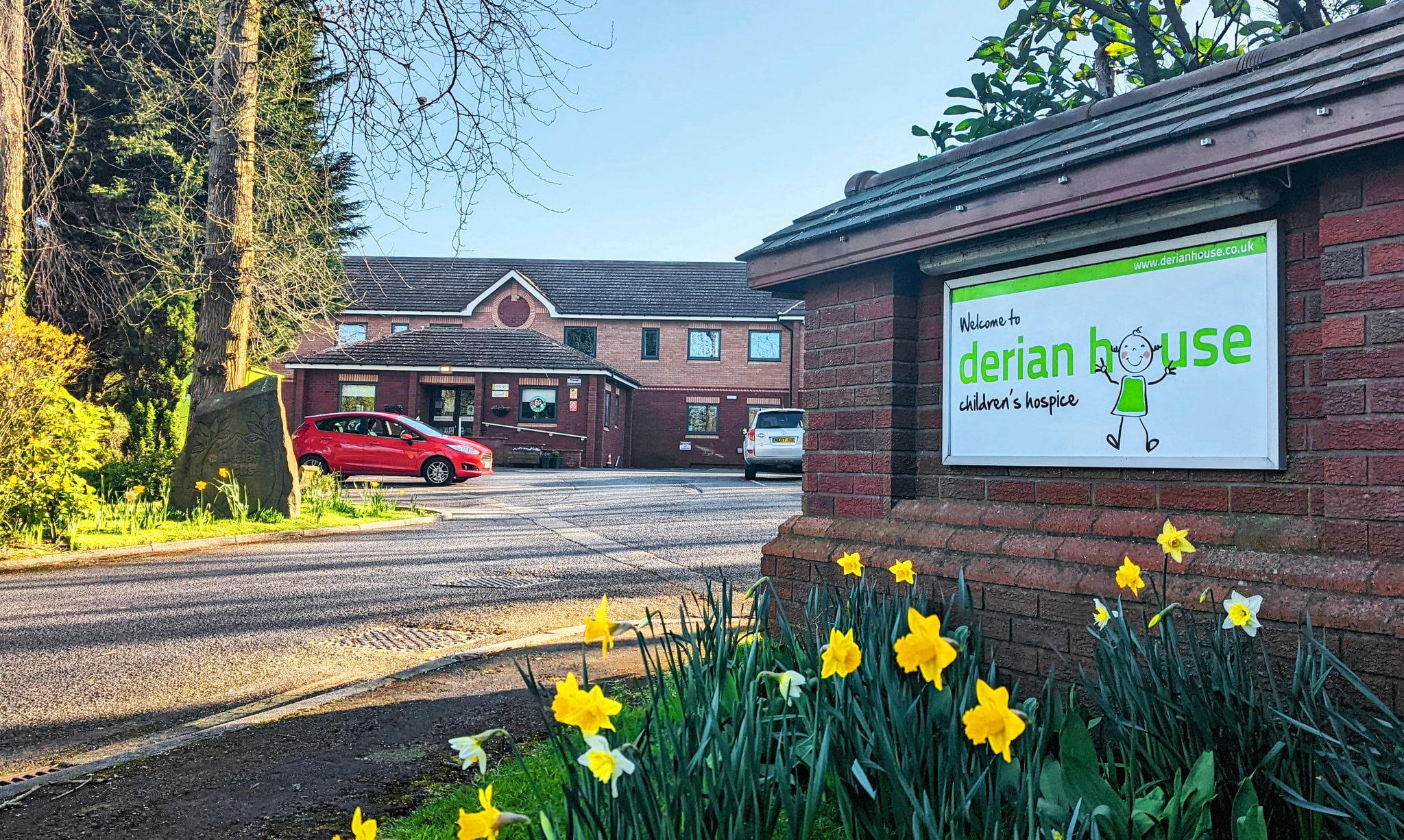 Exterior view of Derian House children's hospice with a large sign on a brick wall, parking lot with cars, and blooming yellow daffodils in the foreground, trees and a building in the background.