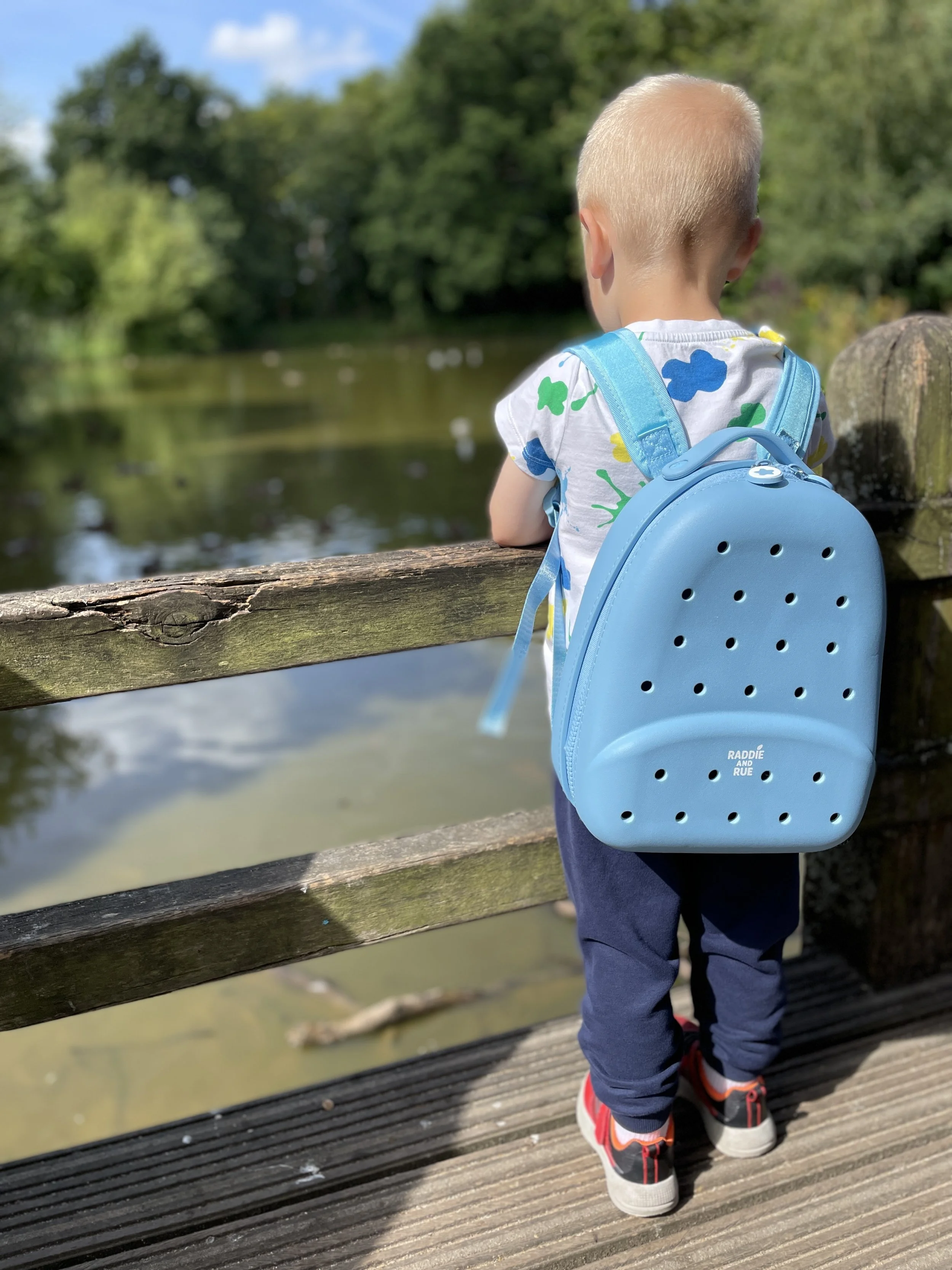 A young child with blonde hair, wearing a blue backpack, a white shirt with colorful patterns, gray pants, and sneakers, stands on a wooden dock looking out at a river with trees in the background.