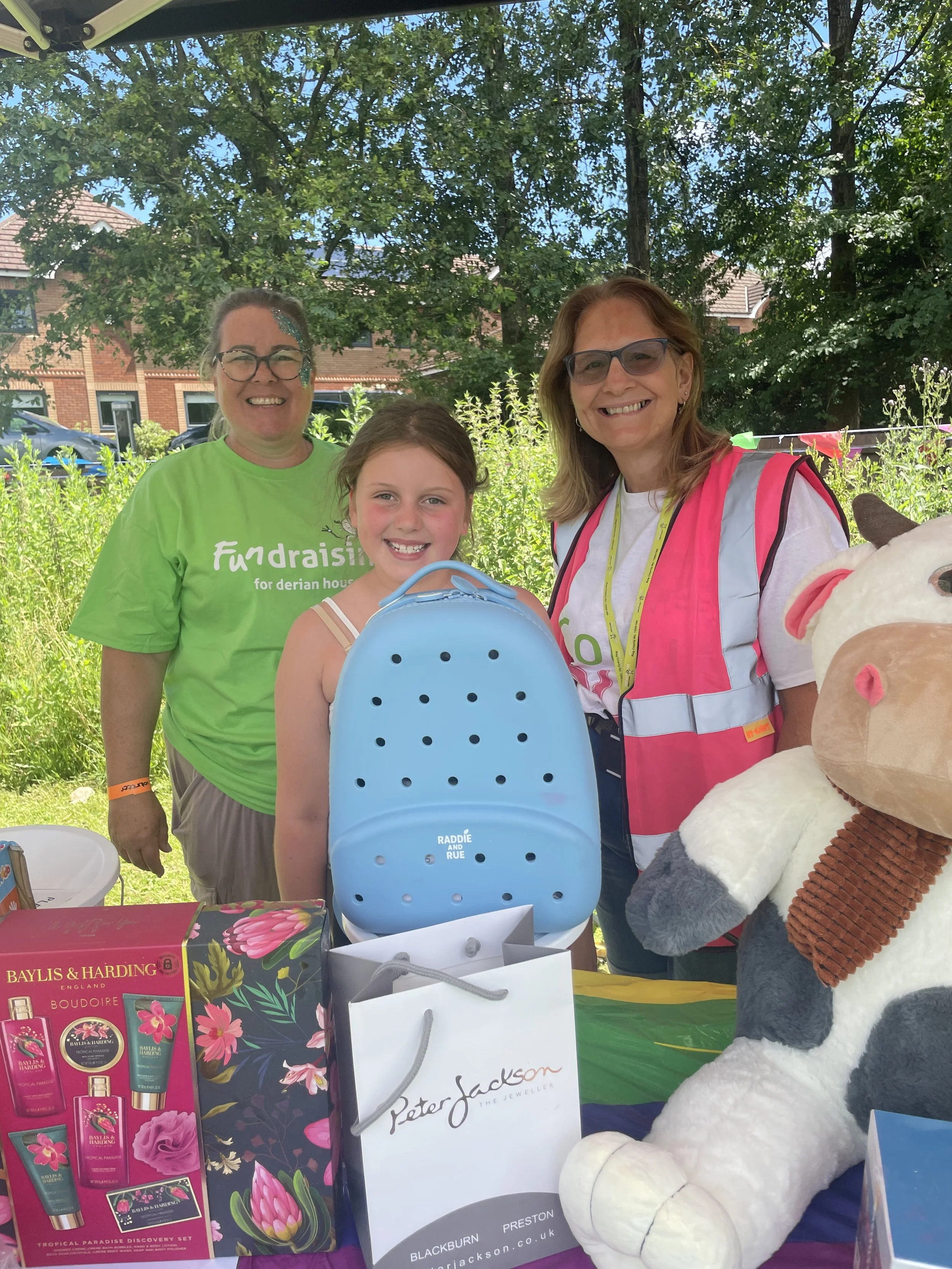 Four smiling people, two women and a young girl, standing outdoors under a tree, with various gift bags and a large teddy bear, at a community event or fair.