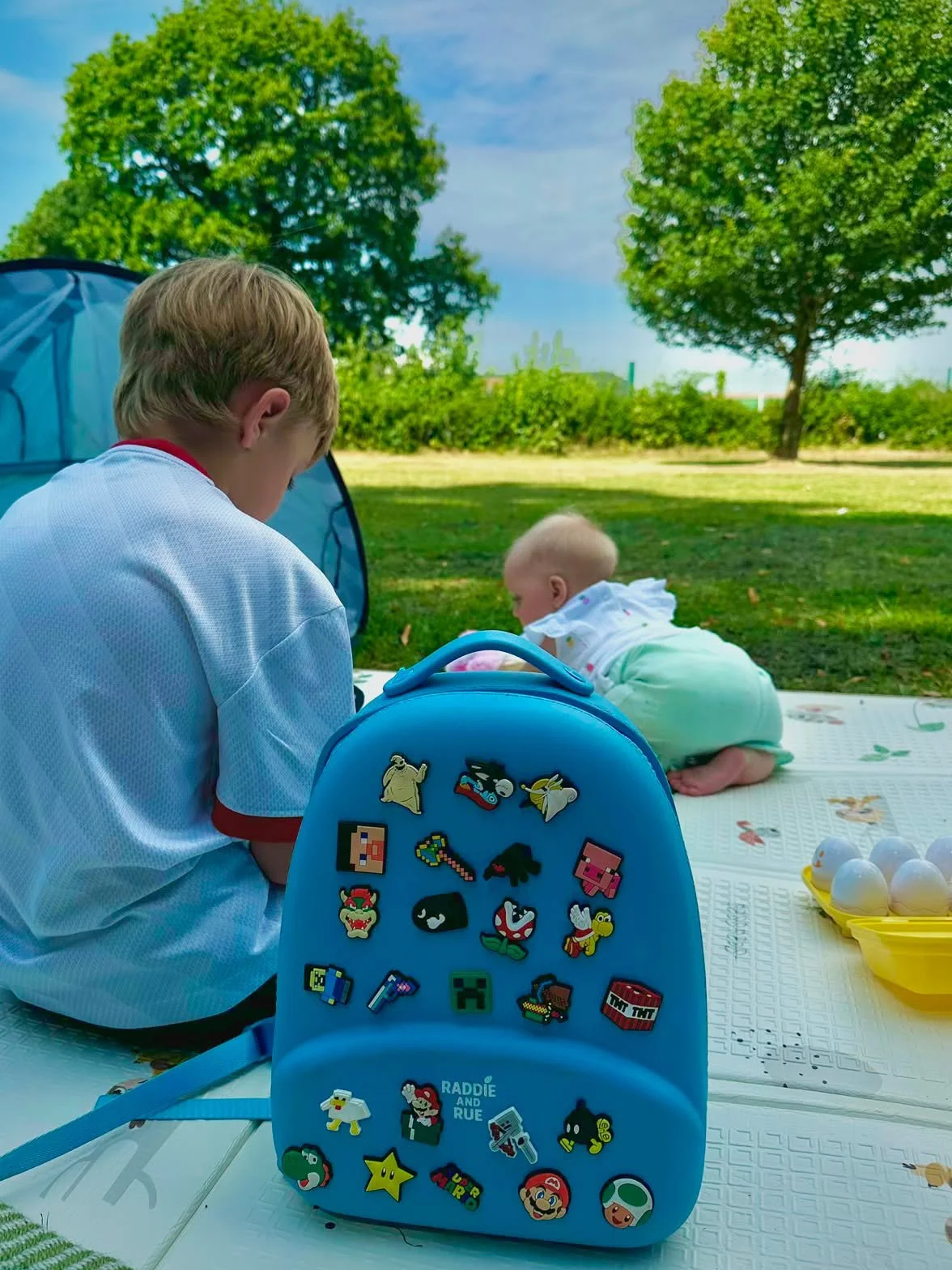 Two children, a boy and a girl, are outdoor on a blanket during daytime, with trees and a blue sky in the background. The boy is sitting with his back to the camera, and the girl is crawling on the blanket. A blue backpack with various colorful stick