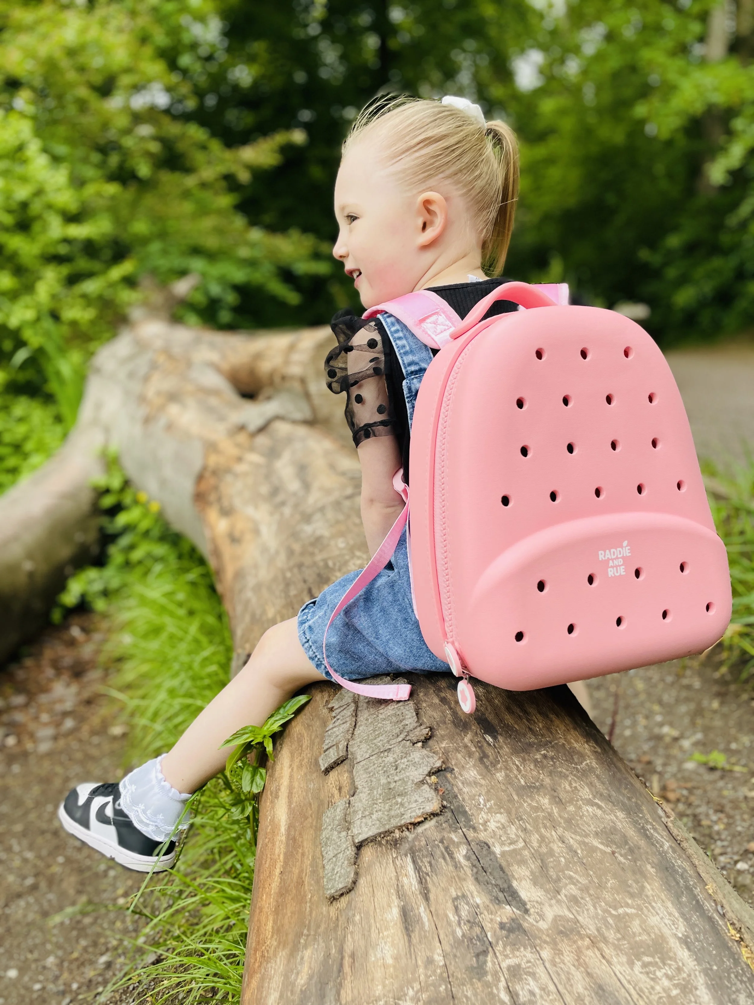 A young girl wearing a black and polka dot top, denim shorts, and grey sneakers with white socks, sitting on a tree branch outdoors, facing sideways with a pink backpack on her back.