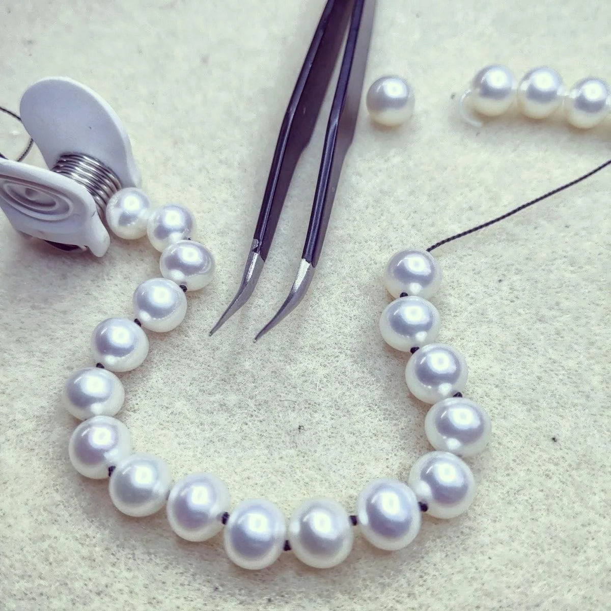 A string of hand-knotted pearls, a pair of tweezers, and jewelry-making tools on a light-colored surface.