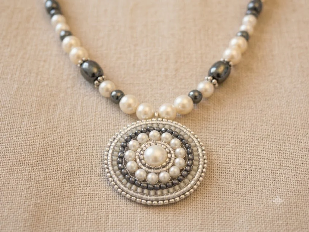 Close-up of a pearl-and-bead necklace design by Lisa Sirlin-Hall, featuring a large concentric-circle pendant on beige fabric.