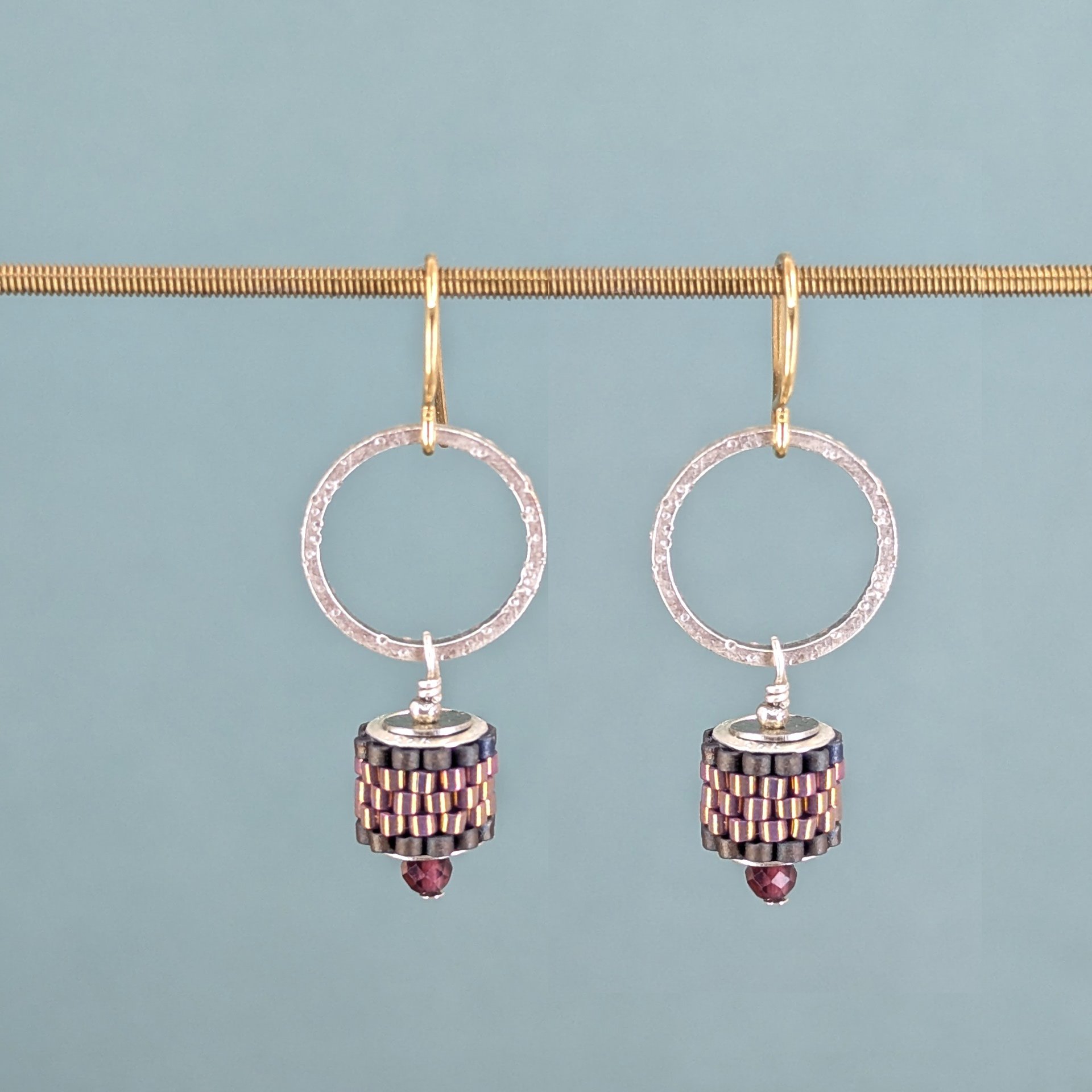 A pair of silver hoop earrings with a textured design, hanging from gold hooks, featuring Japanese glass and red gemstones, against a blue background.