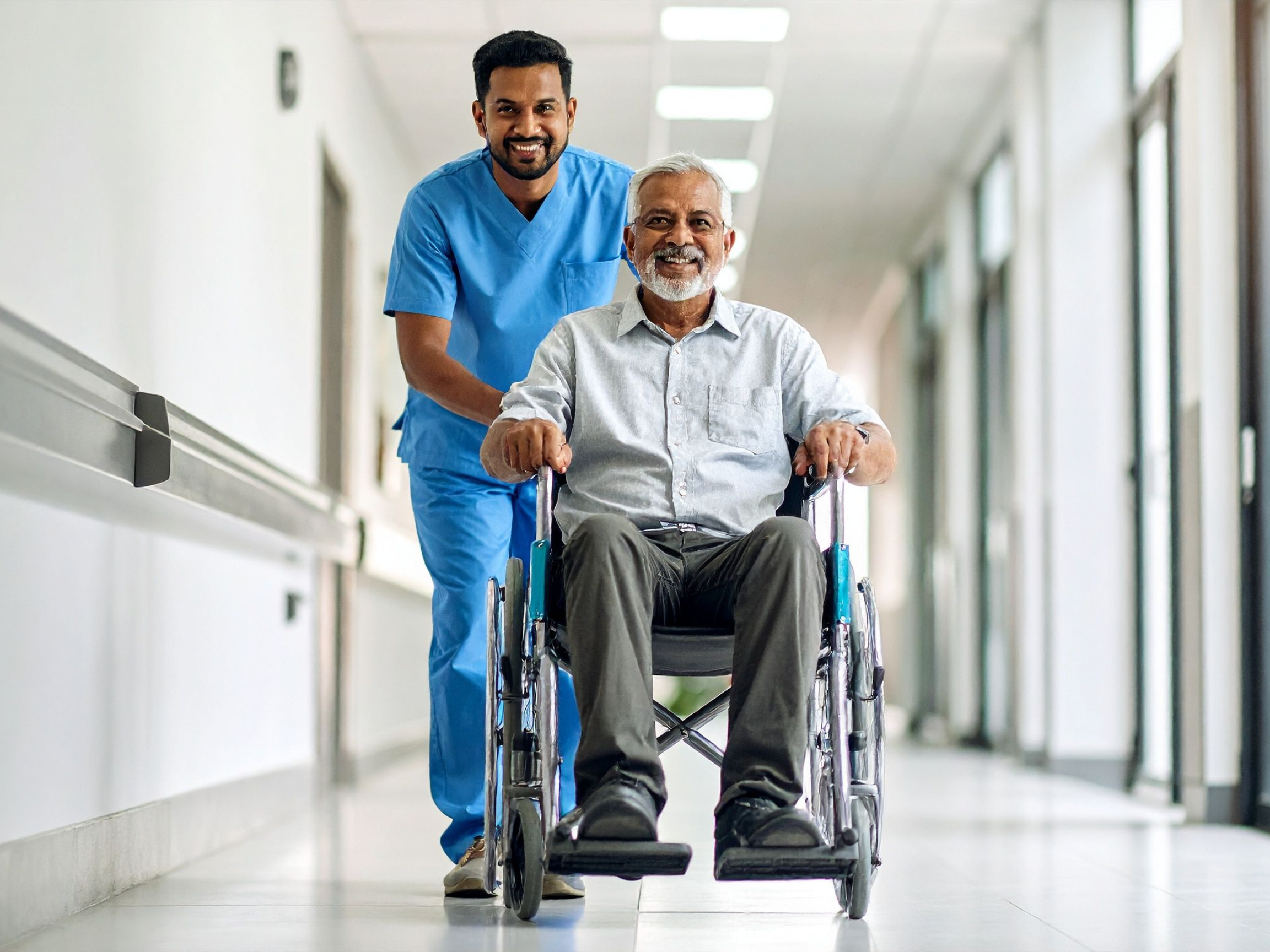 A healthcare worker pushing an elderly man in a wheelchair down a hospital corridor, both smiling.