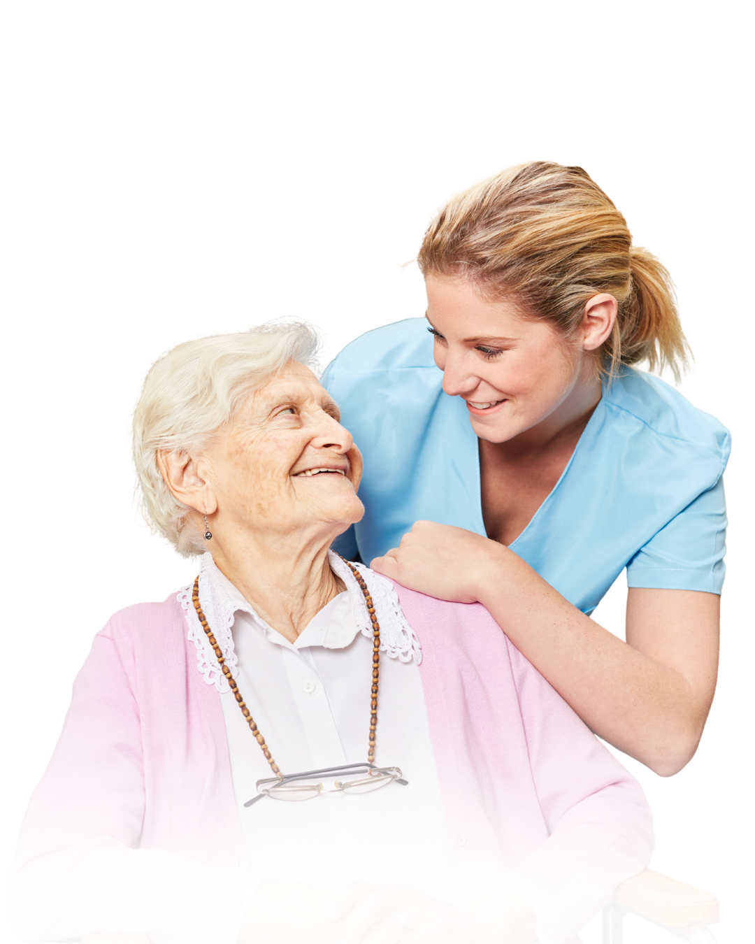A young female caregiver with light brown hair smiling and leaning toward an elderly woman with white hair, who is wearing glasses and a pink cardigan, as they make eye contact and share a joyful moment.