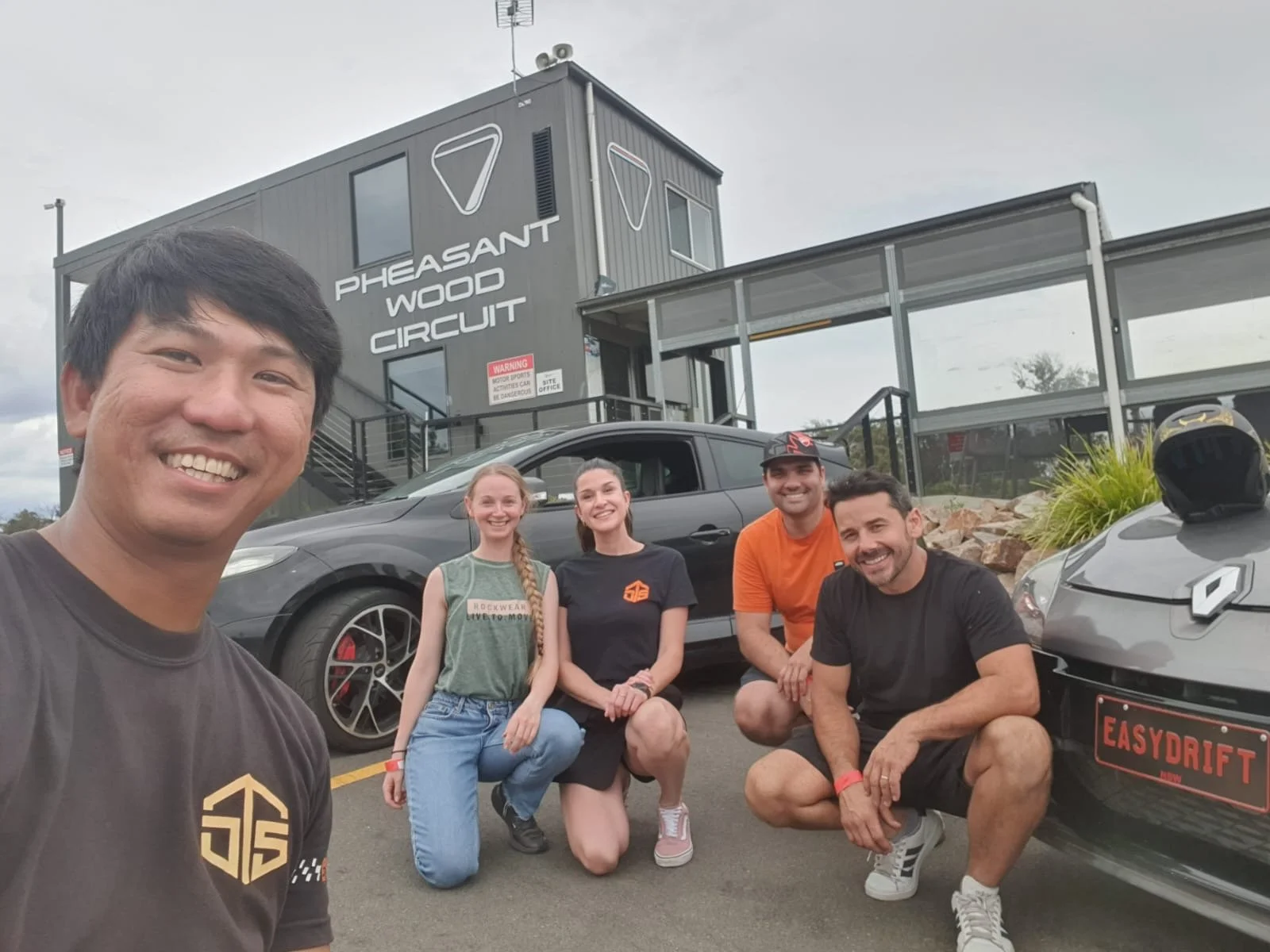 A group of five people taking a selfie in front of the Pheasant Wood Circuit race track, with cars behind them, including a gray sports car with a race helmet on its hood, at a professional racing venue.