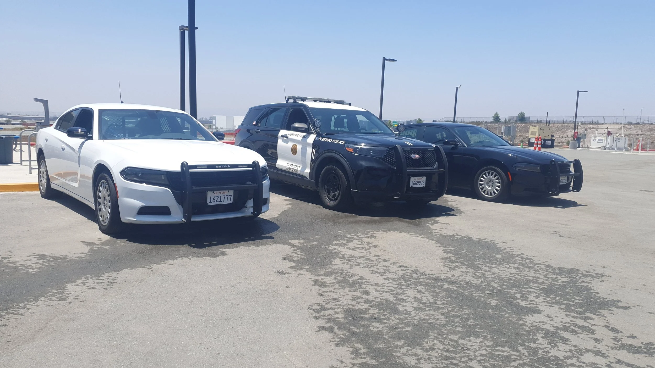 Three police cars parked in a parking lot, including a white sedan, a black police SUV, and a black police sedan, with a clear sky and distant industrial structures in the background.