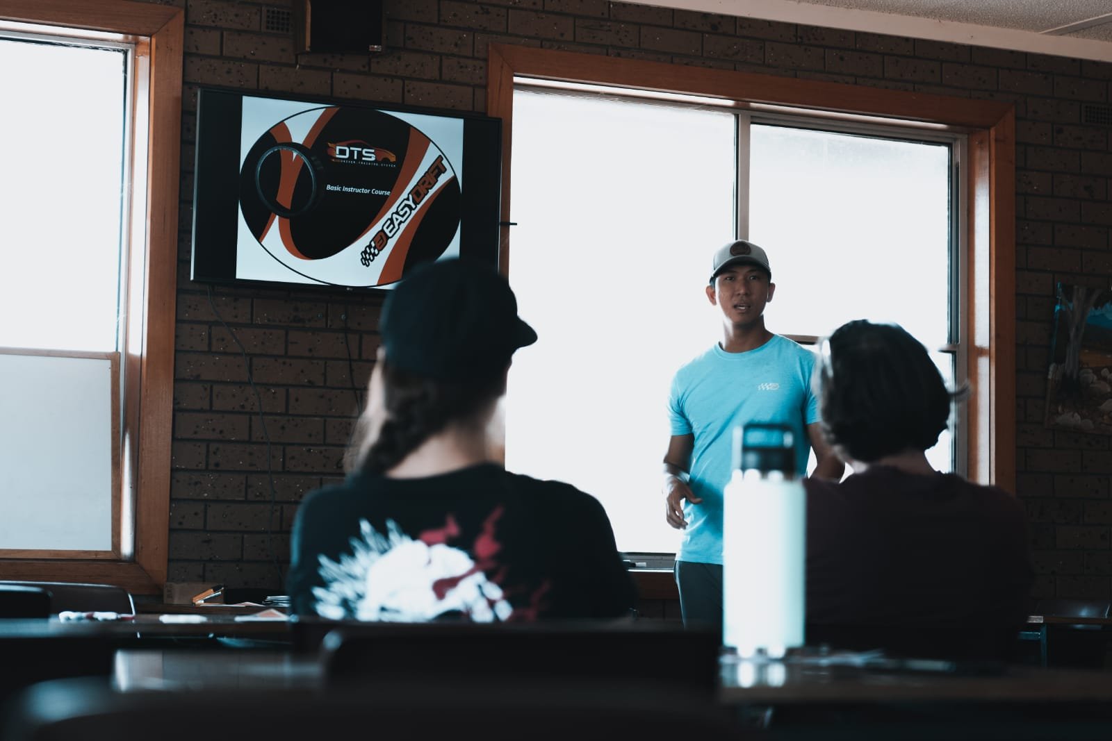 Instructor standing at the front of a classroom with two seated students, a monitor displaying a presentation behind him, and large windows in the background. Theory about Easydrift.