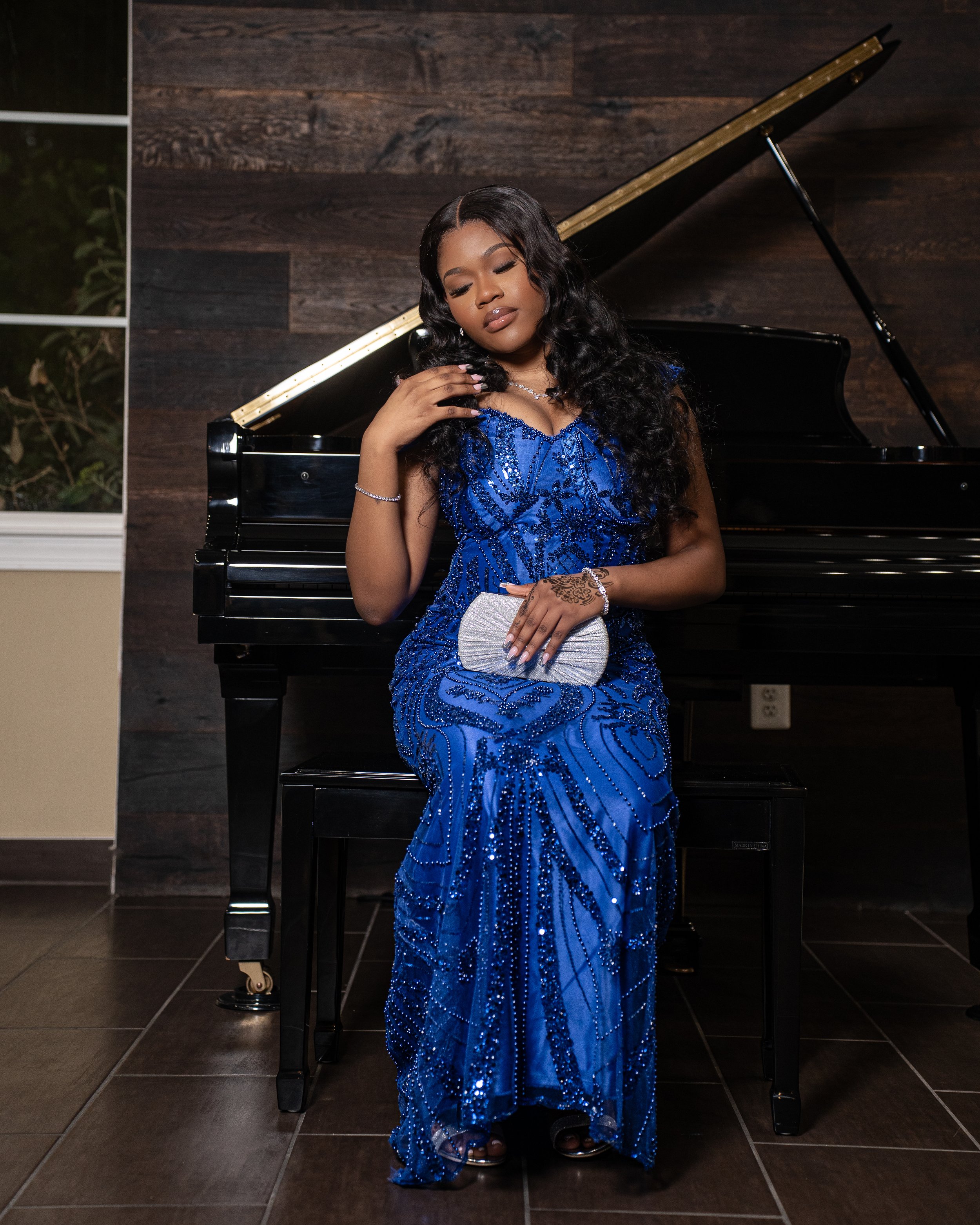 A young woman in a sparkling blue evening gown sitting on a piano bench next to a grand piano, holding a silver clutch, with long dark hair and wearing jewelry, posing with eyes closed.
