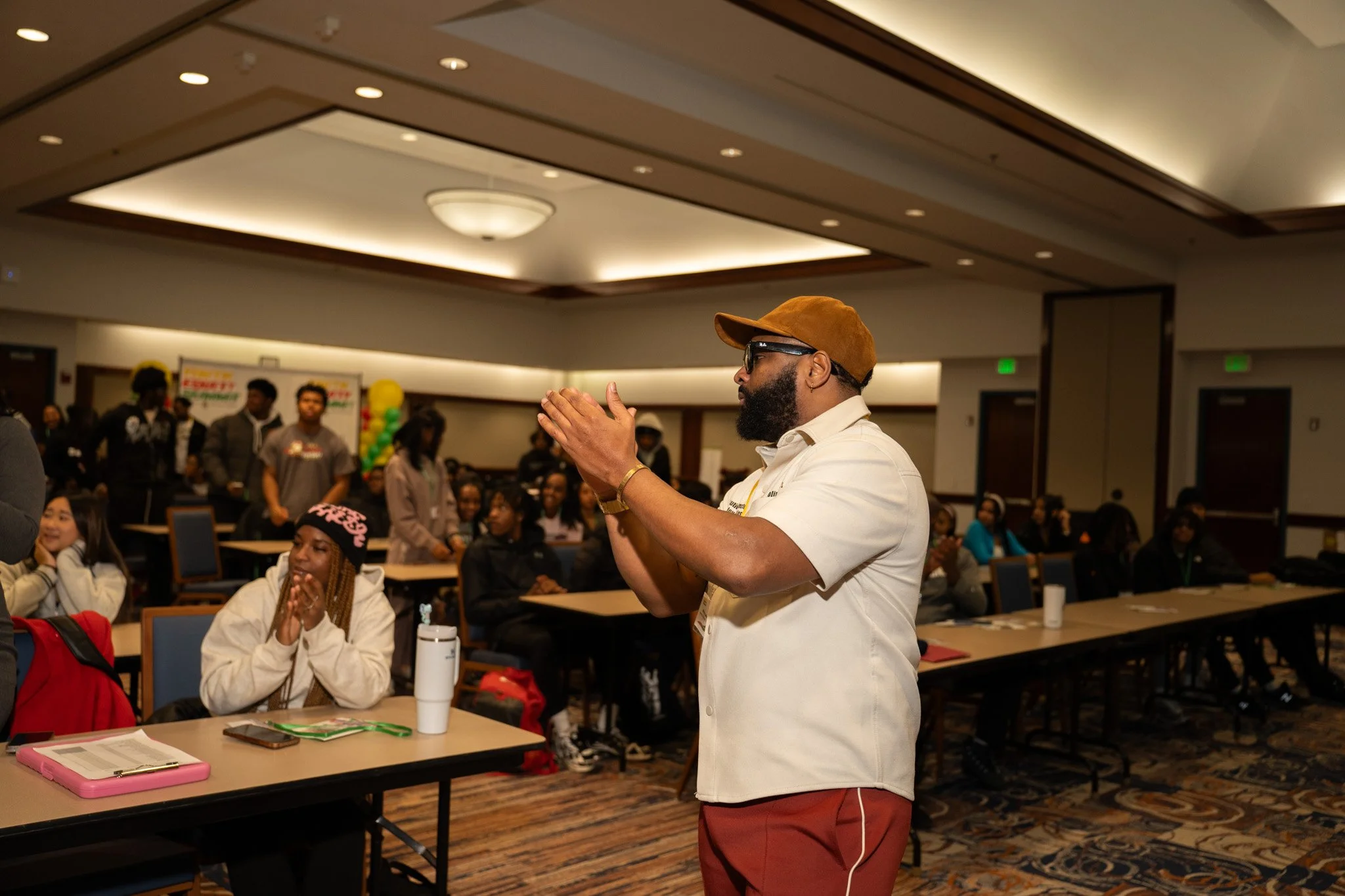 A man with a beard, wearing sunglasses, a brown hat, a white shirt, and brown pants, stands and claps in a conference or seminar room filled with seated attendees, some smiling and looking towards him.