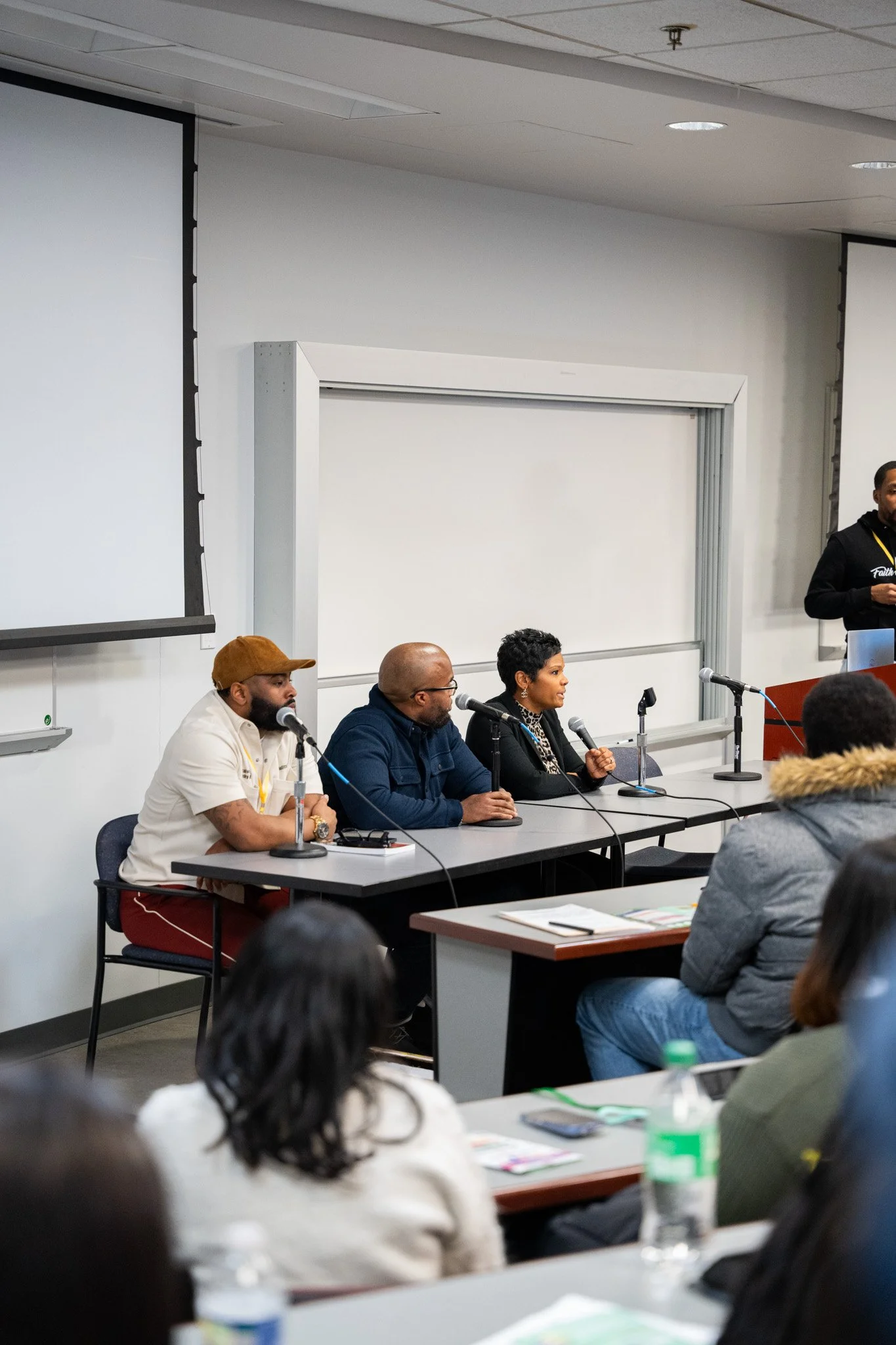 Panel of three people sitting at a table with microphones in front of a classroom or conference room, with an audience observing.
