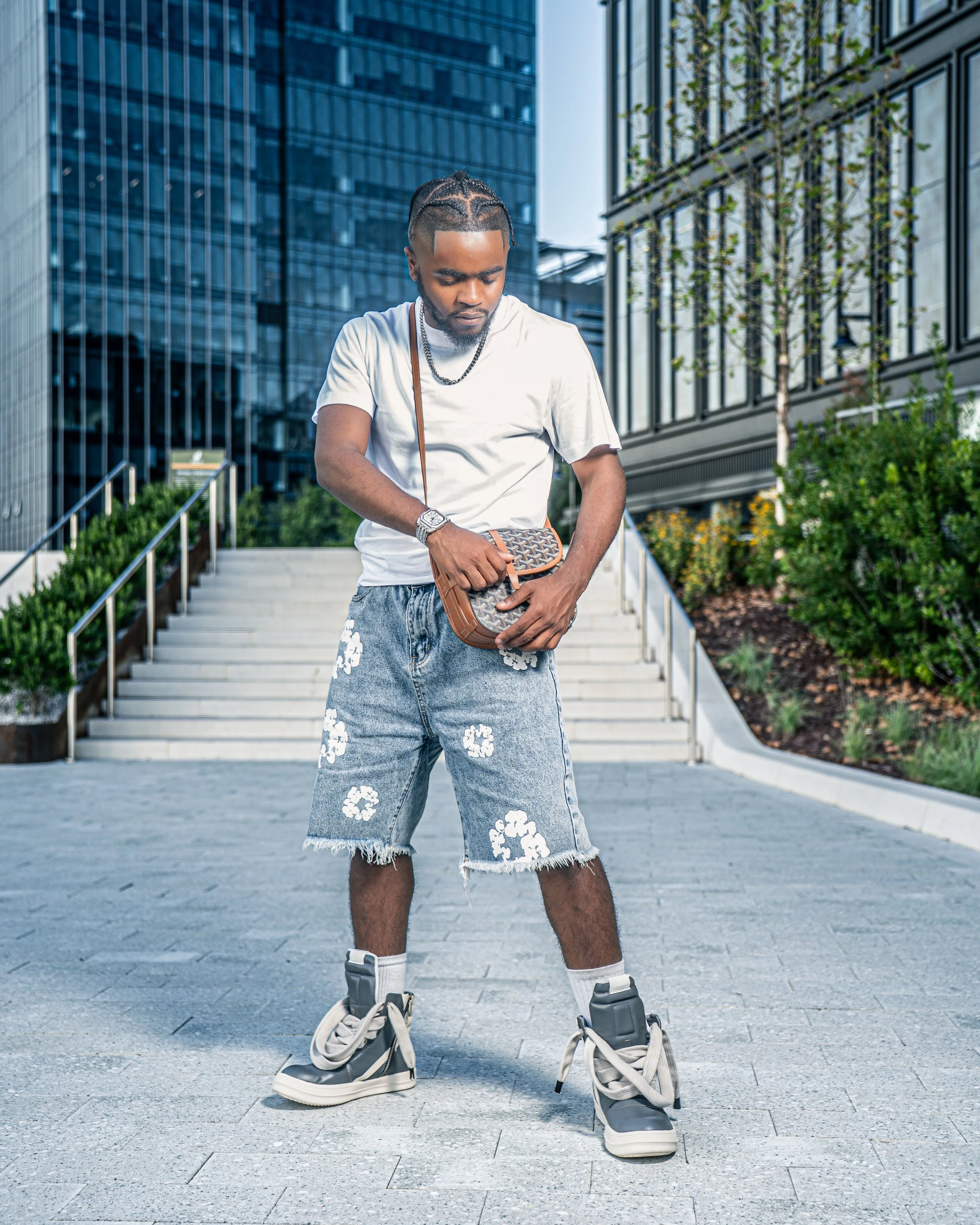 Young man standing outdoors in an urban area, looking down, wearing a white t-shirt, distressed floral denim shorts, white socks, and high-top roller skates, holding a small patterned bag, with modern glass buildings and stairs in the background.
