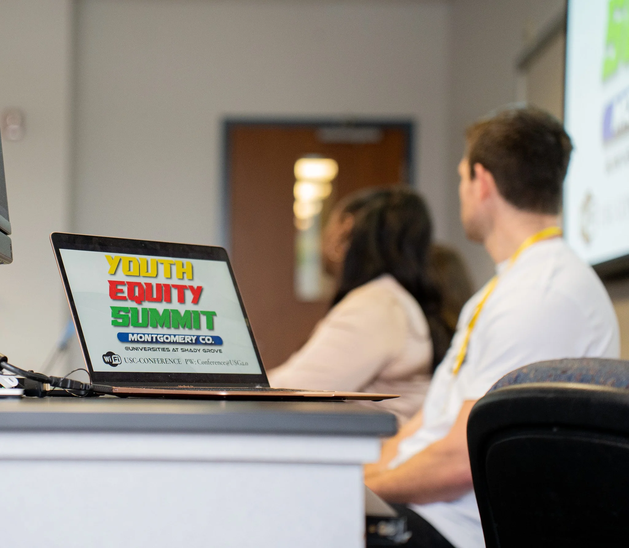 A conference room with a laptop displaying a colorful sign for the Youth Equity Summit in Montgomery County and several attendees in focus, sitting and listening.
