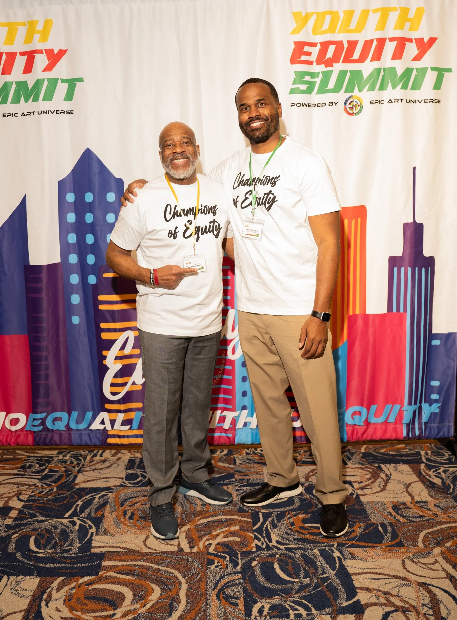 Two men standing together at the Youth Equity Summit, both smiling and wearing white T-shirts that read 'Champions of Equity.' They are posing in front of a colorful backdrop with the event's name and cityscape graphics.