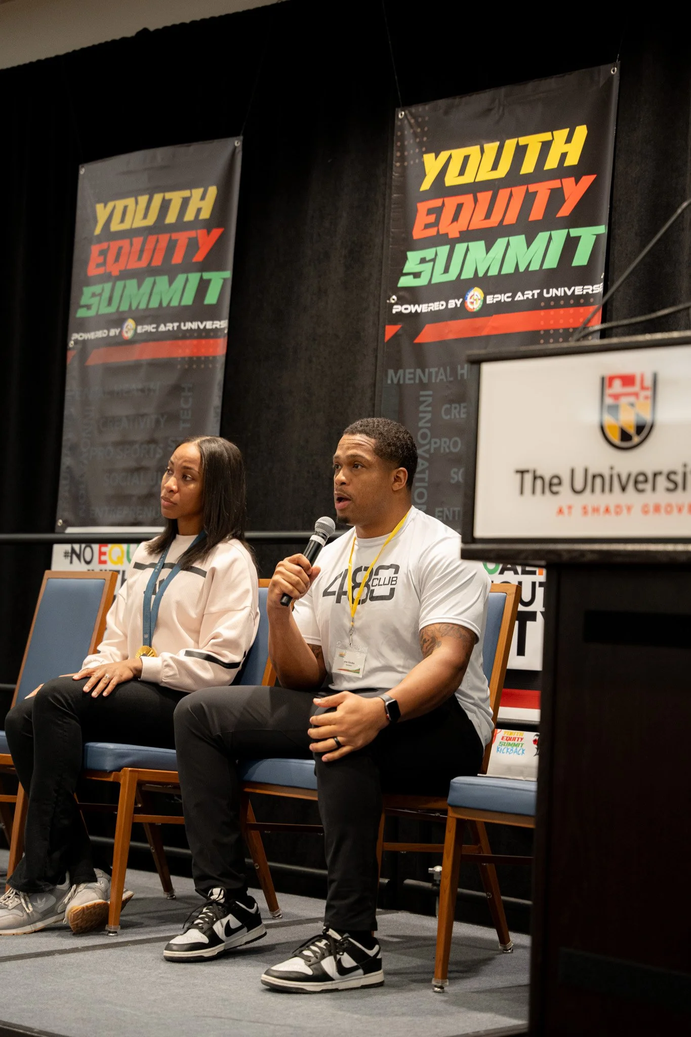 Two people sitting on stage at the Youth Equity Summit, with one man speaking into a microphone. The background features banners with the event name and logos.