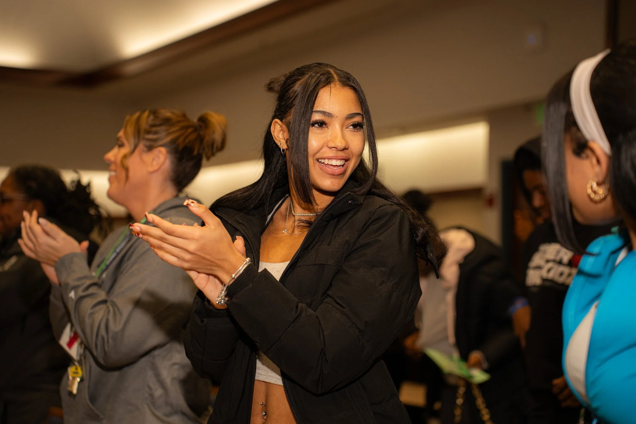 A group of women standing in a room, clapping and smiling, with one woman in the foreground wearing a black jacket, a crop top, and jewelry.